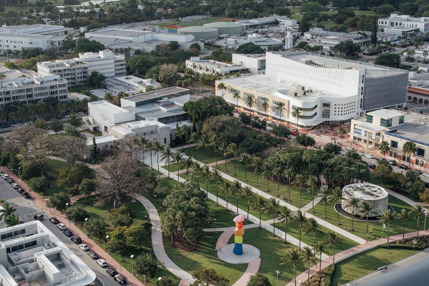 Aerial view of a urban park with winding paths, palm trees, colorful sculpture, and modern white buildings in Miami.