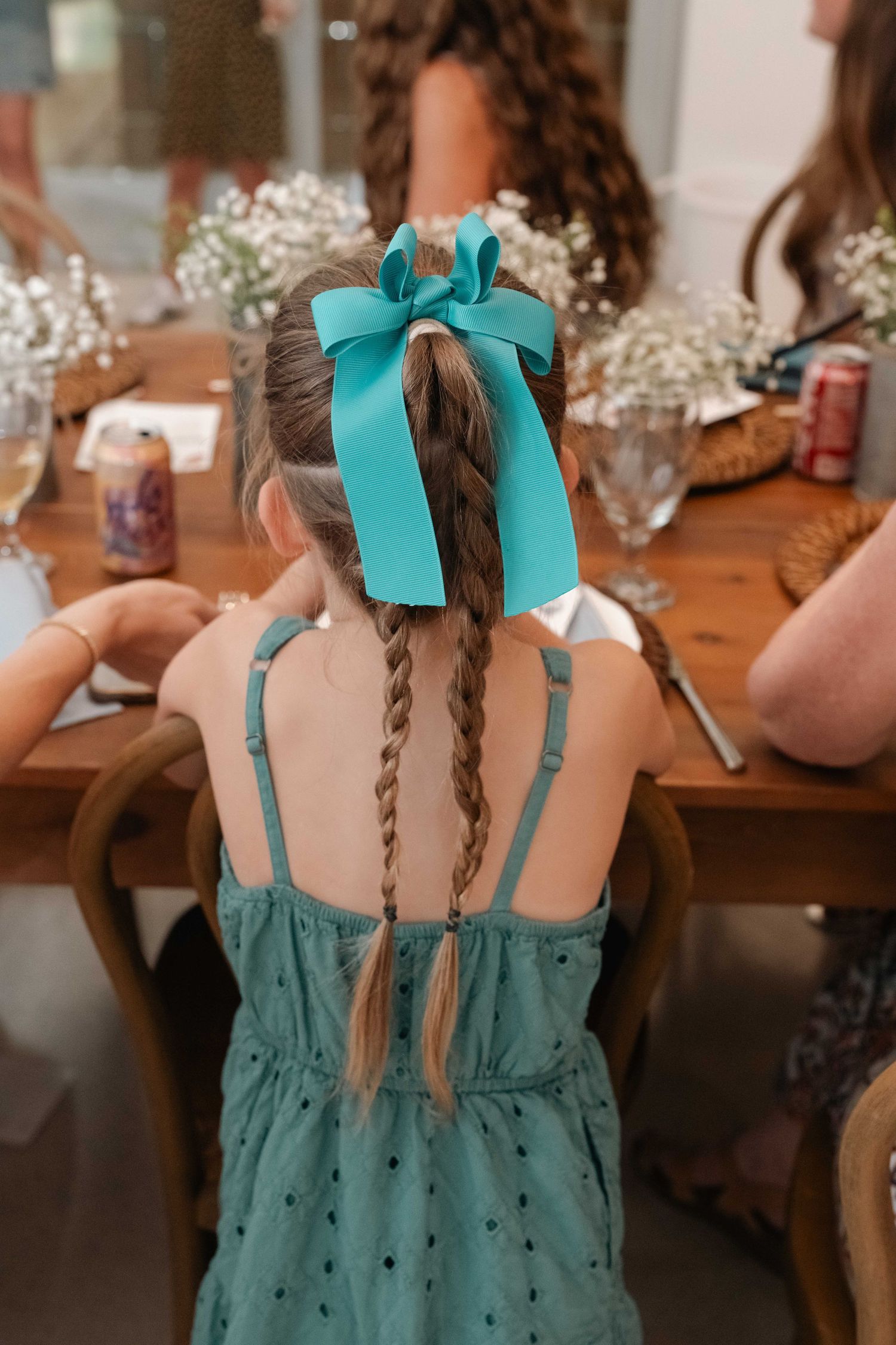 Young girl with braided pigtails and teal ribbon bow sitting at a wooden dining table.