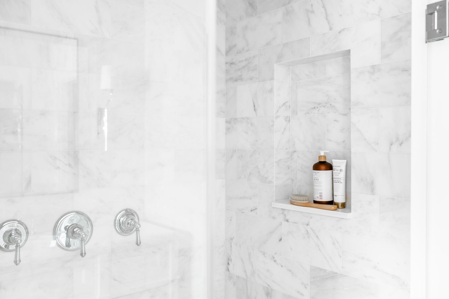 White marble shower with built-in niche shelf holding amber bottle and wood tray, chrome fixtures visible.