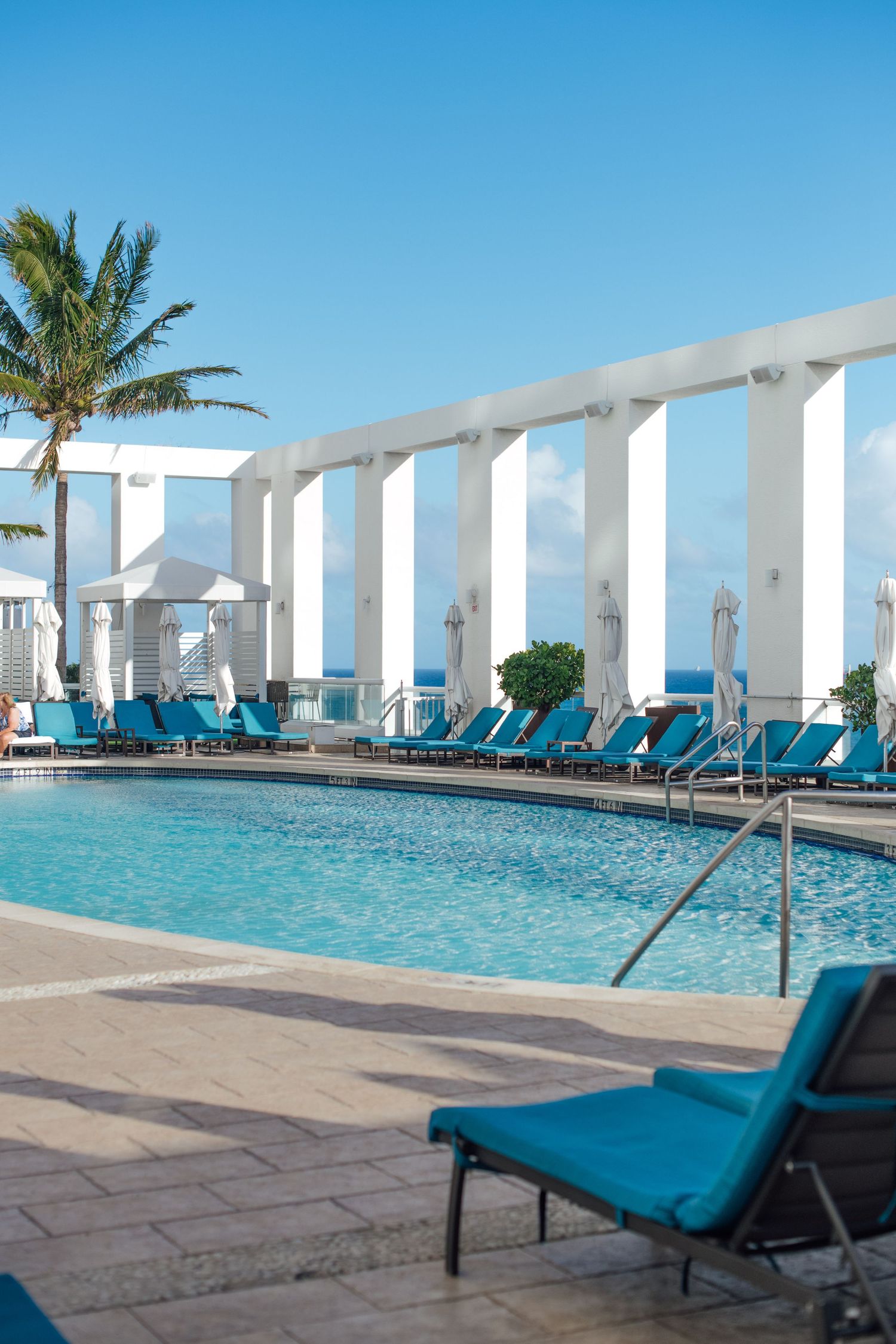 Resort pool with turquoise lounge chairs, tall white columns, palm tree, and ocean view on sunny day.