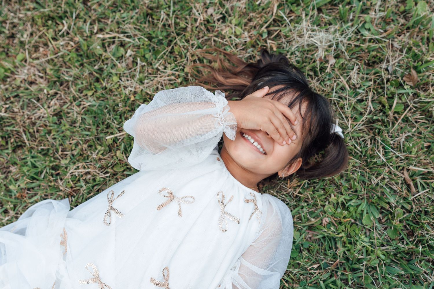 Young girl in white dress lying on grass, laughing joyfully with hands covering her eyes.