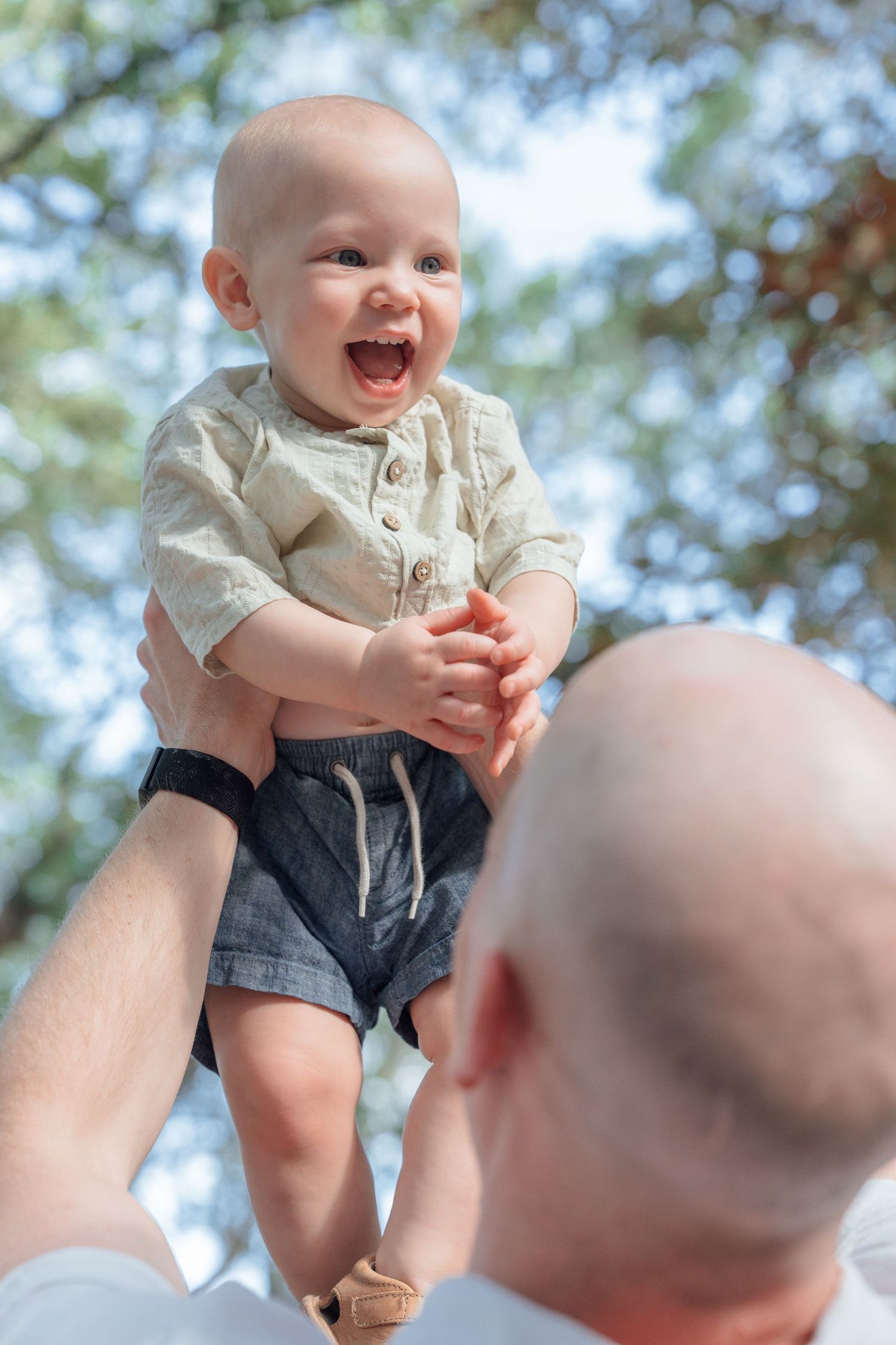 Joyful baby laughing while being lifted up by a bald father outdoors in a sunny park setting.