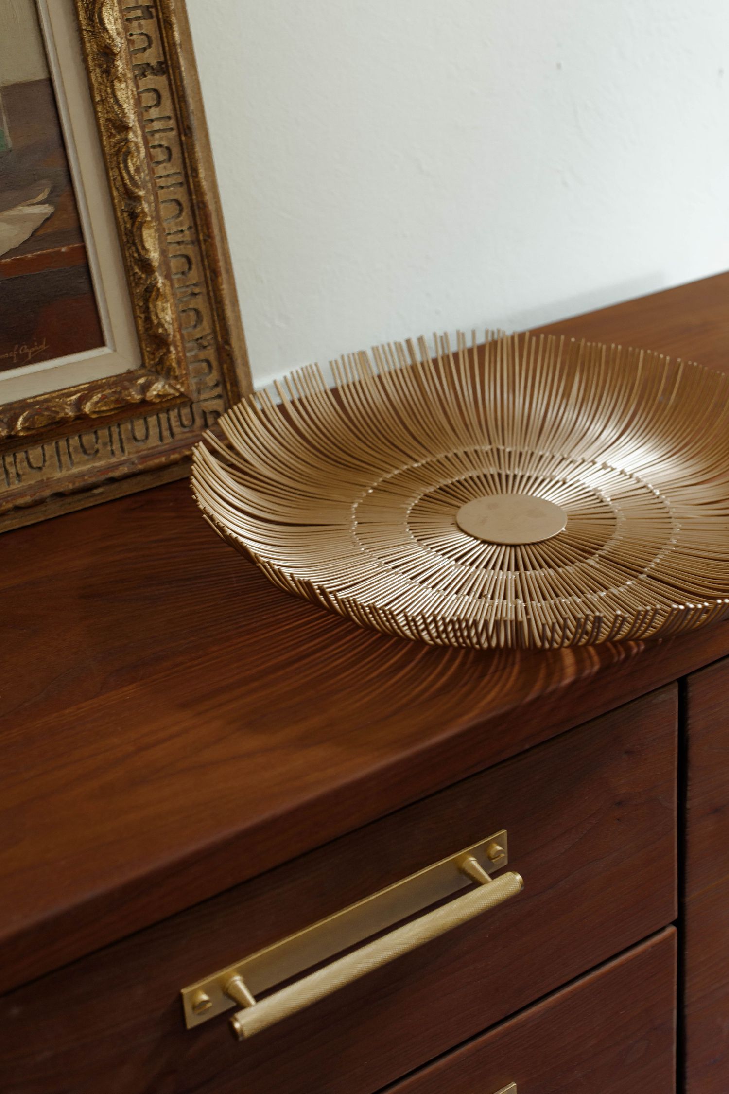 Gold sunburst decorative plate on a walnut wood dresser with brass hardware, beside an ornate gold framed artwork.