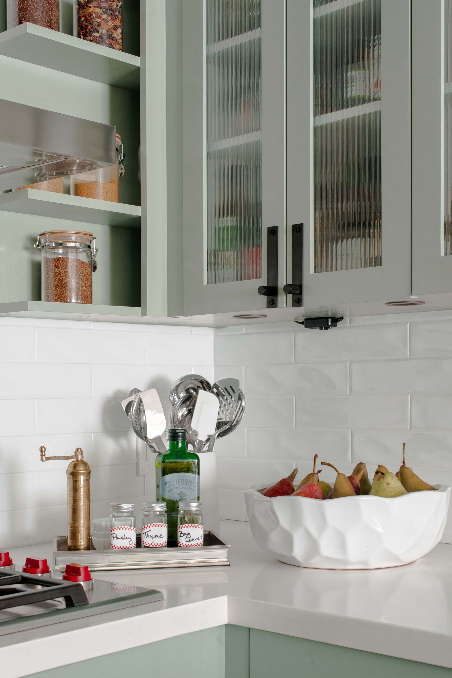 Sage green kitchen cabinets with glass doors, white subway tile backsplash, and a white bowl of pears on the countertop.