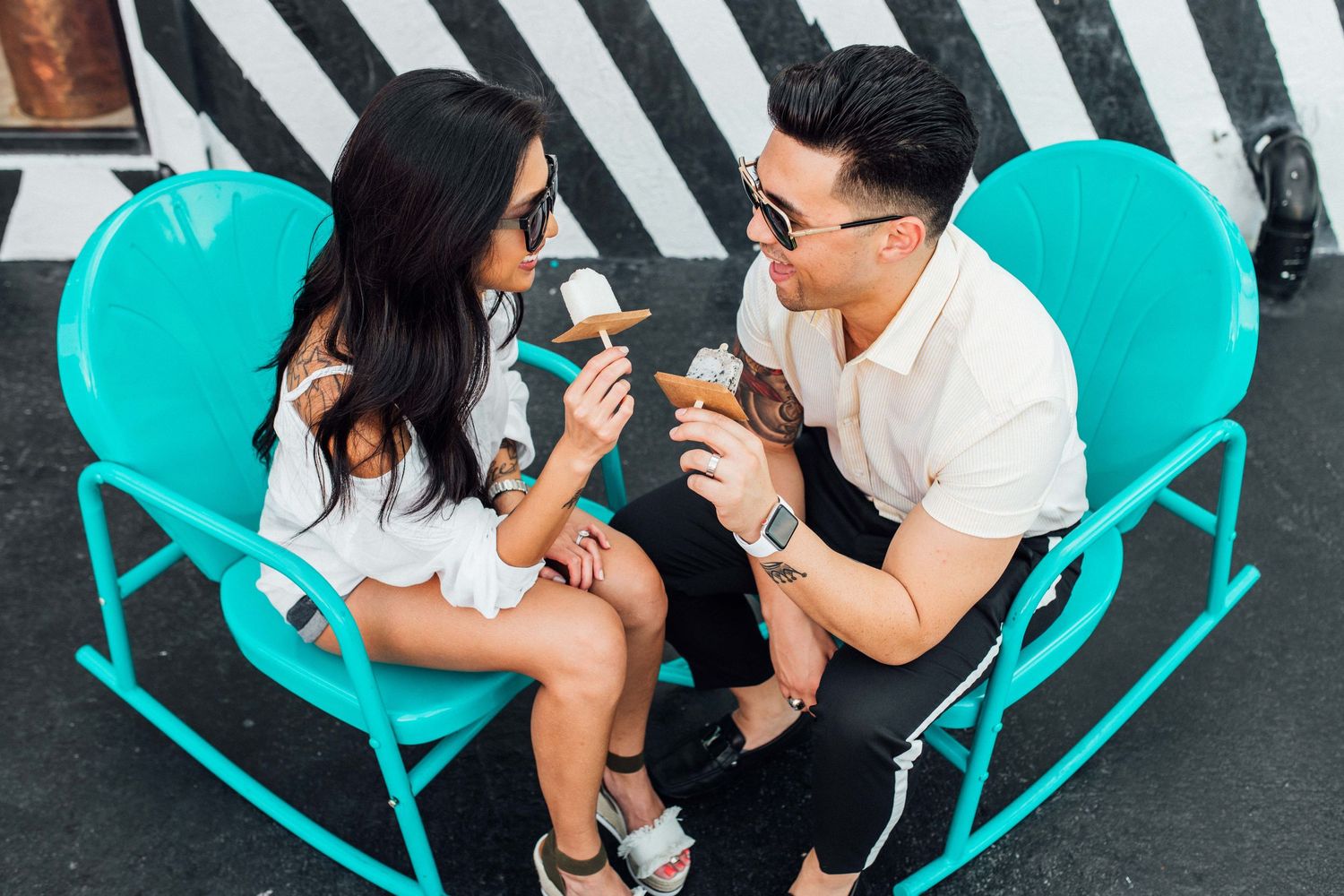 Couple sharing ice cream cones while sitting on teal rocking chairs outdoors.