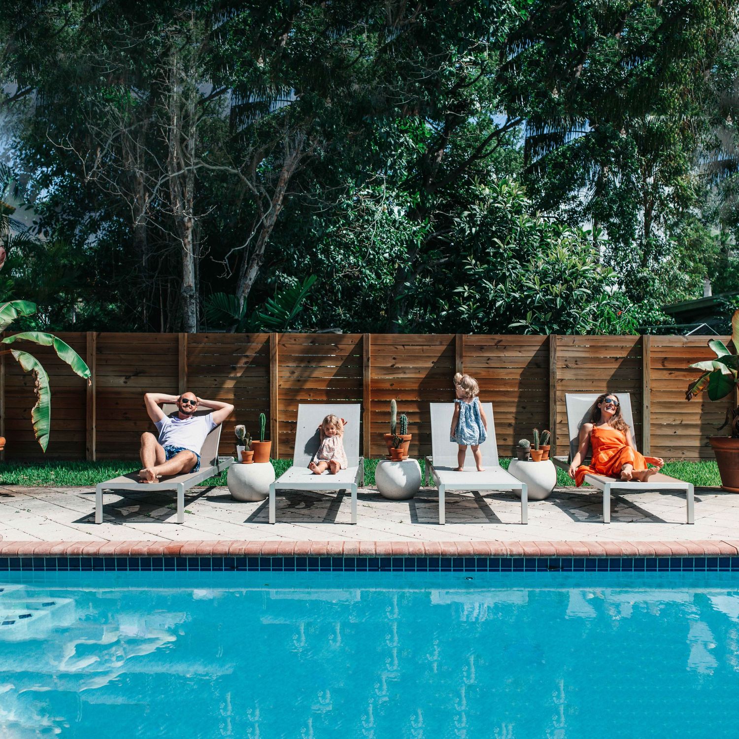Three women relaxing on lounge chairs beside a blue swimming pool surrounded by lush greenery.