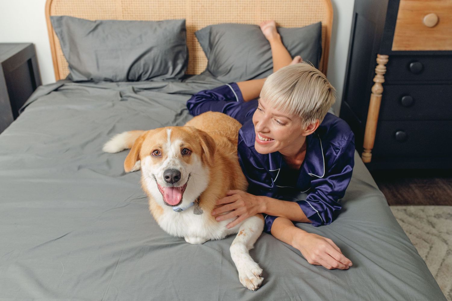 Smiling blonde woman cuddling with a happy golden dog on gray bed sheets.