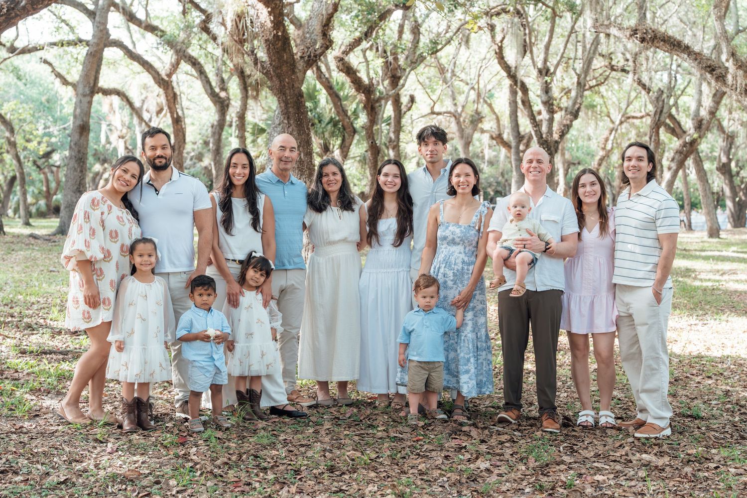 Large family group portrait outdoors under moss-covered oak trees, wearing coordinated light blue and white outfits.
