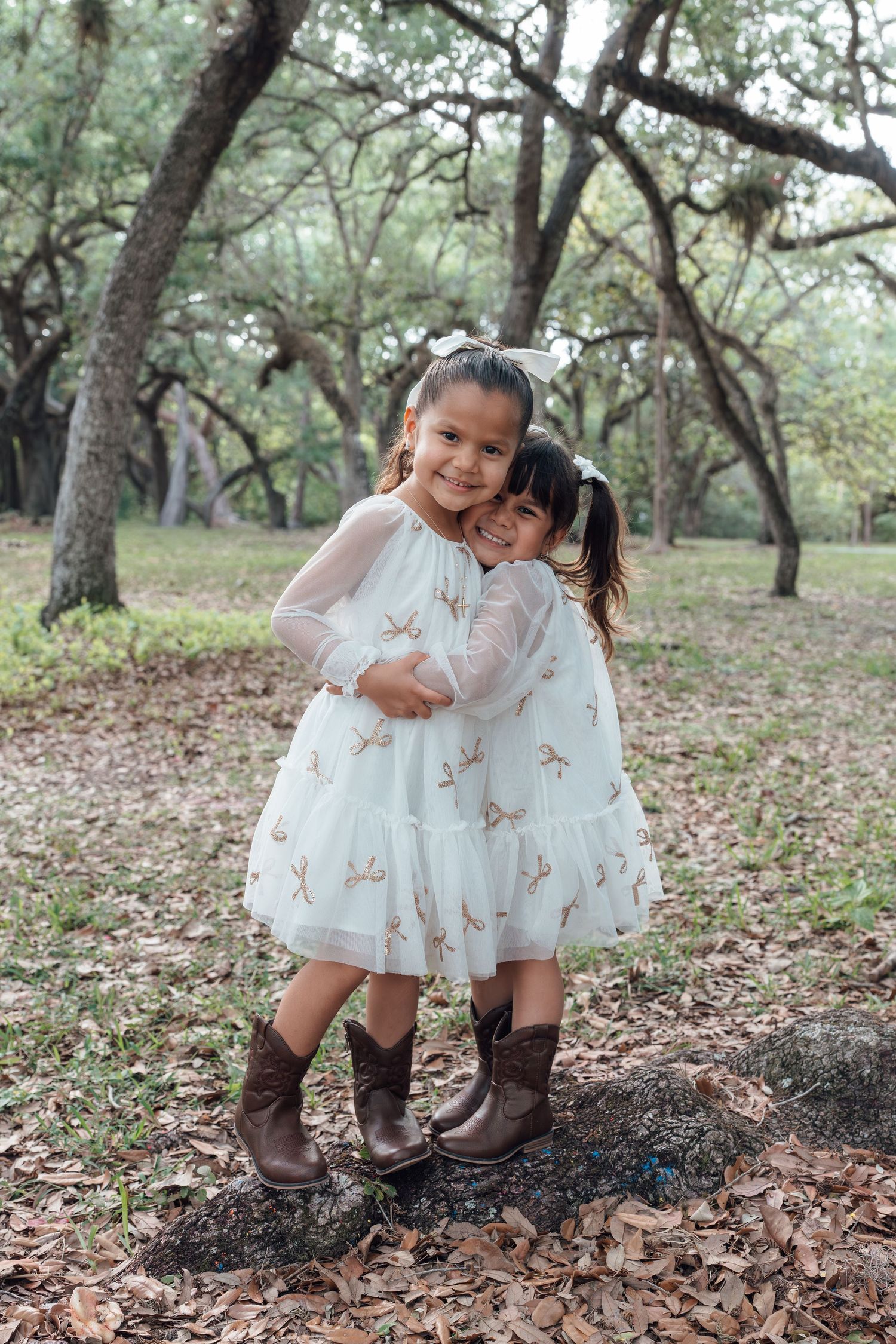 Two young girls in white tulle dresses and brown cowboy boots hugging outdoors among trees covered in Spanish moss.