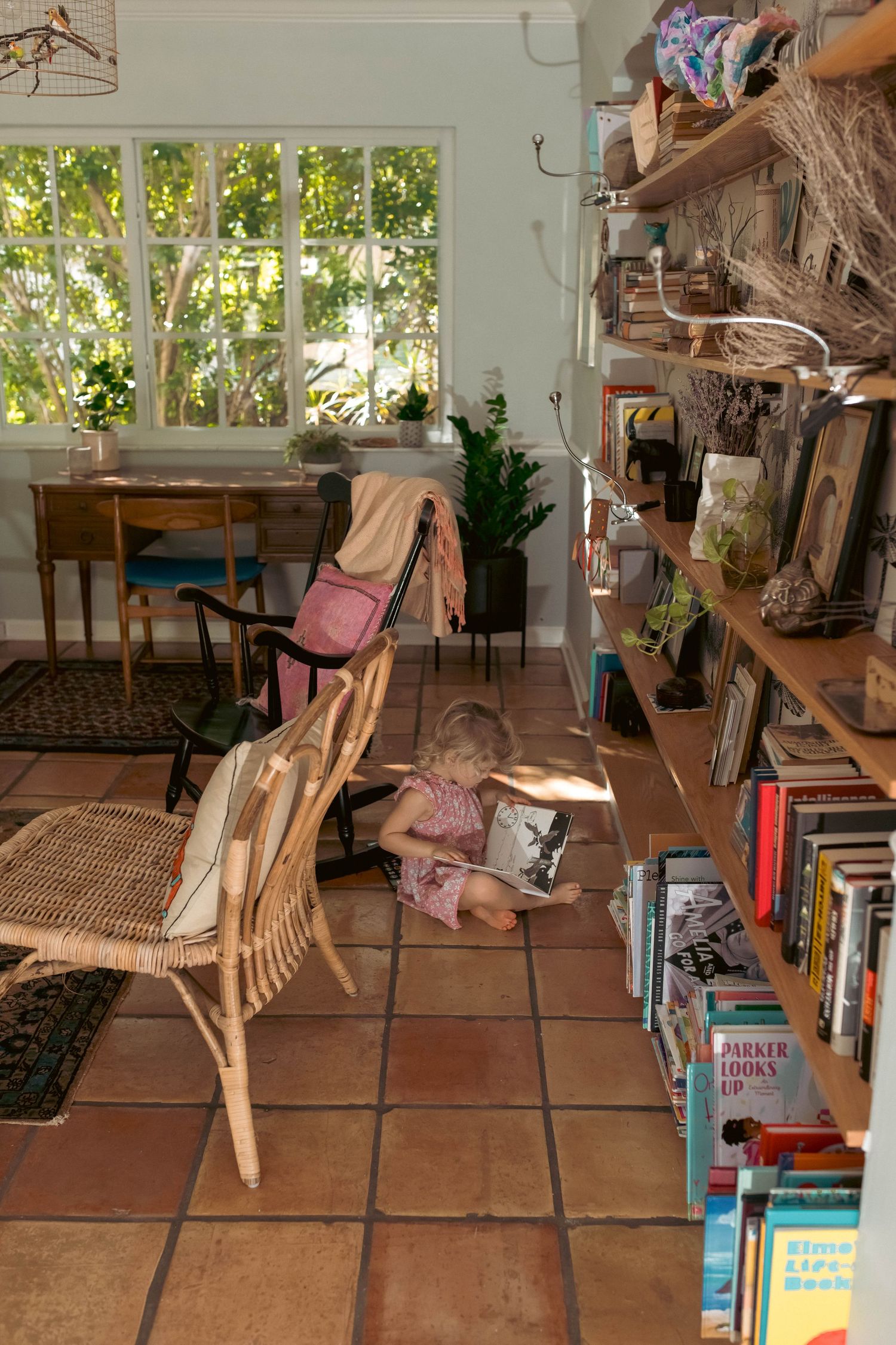 Young child reading a book on the floor of a cozy sunlit home library with wooden shelves.