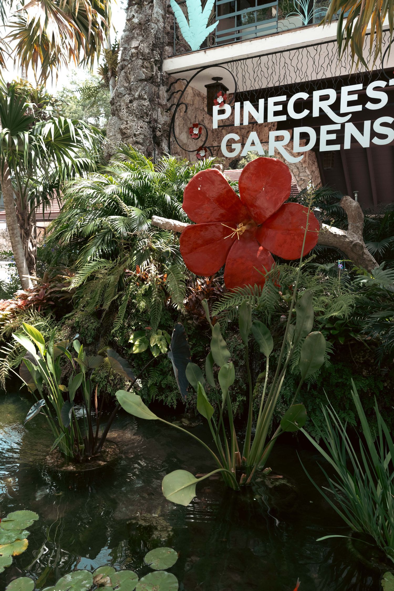 Pinecrest Gardens entrance sign surrounded by lush tropical plants and a large red hibiscus sculpture.