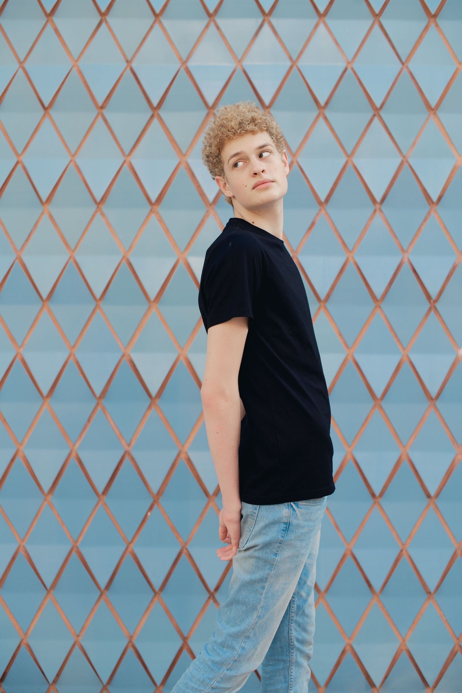 Young man with curly blonde hair in black t-shirt and jeans posing against geometric wall.