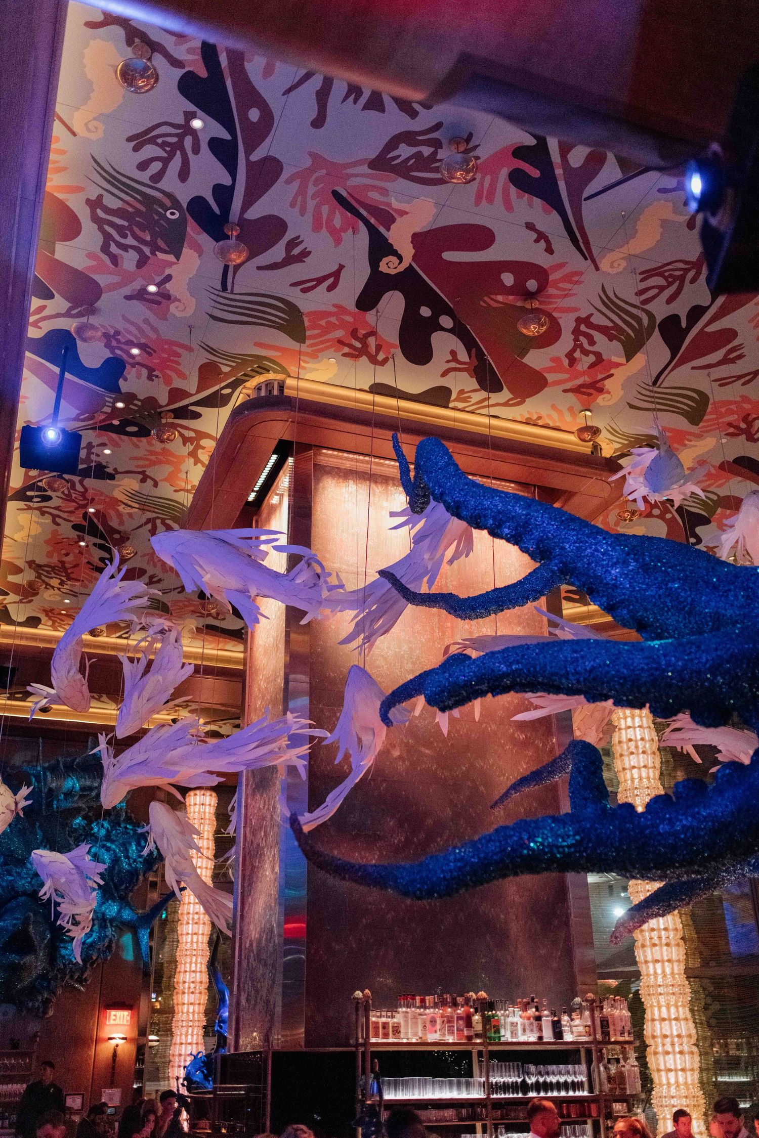 Dramatic restaurant interior with artistic ceiling murals, blue glittering tree branches, and jellyfish decorations above a bar.