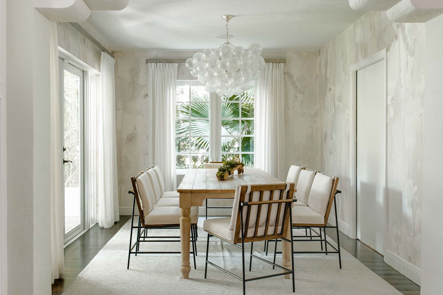 Elegant dining room with marble walls, natural wood table, upholstered chairs, white rug, and a floral chandelier.
