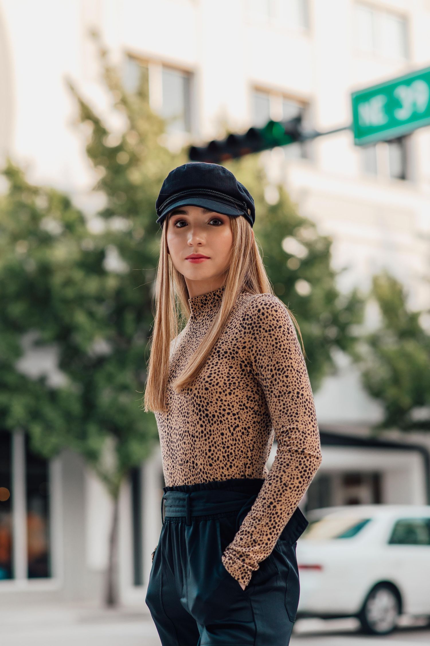 Trendy blonde woman in leopard bodysuit and black leather pants wearing a newsboy cap on a city street.