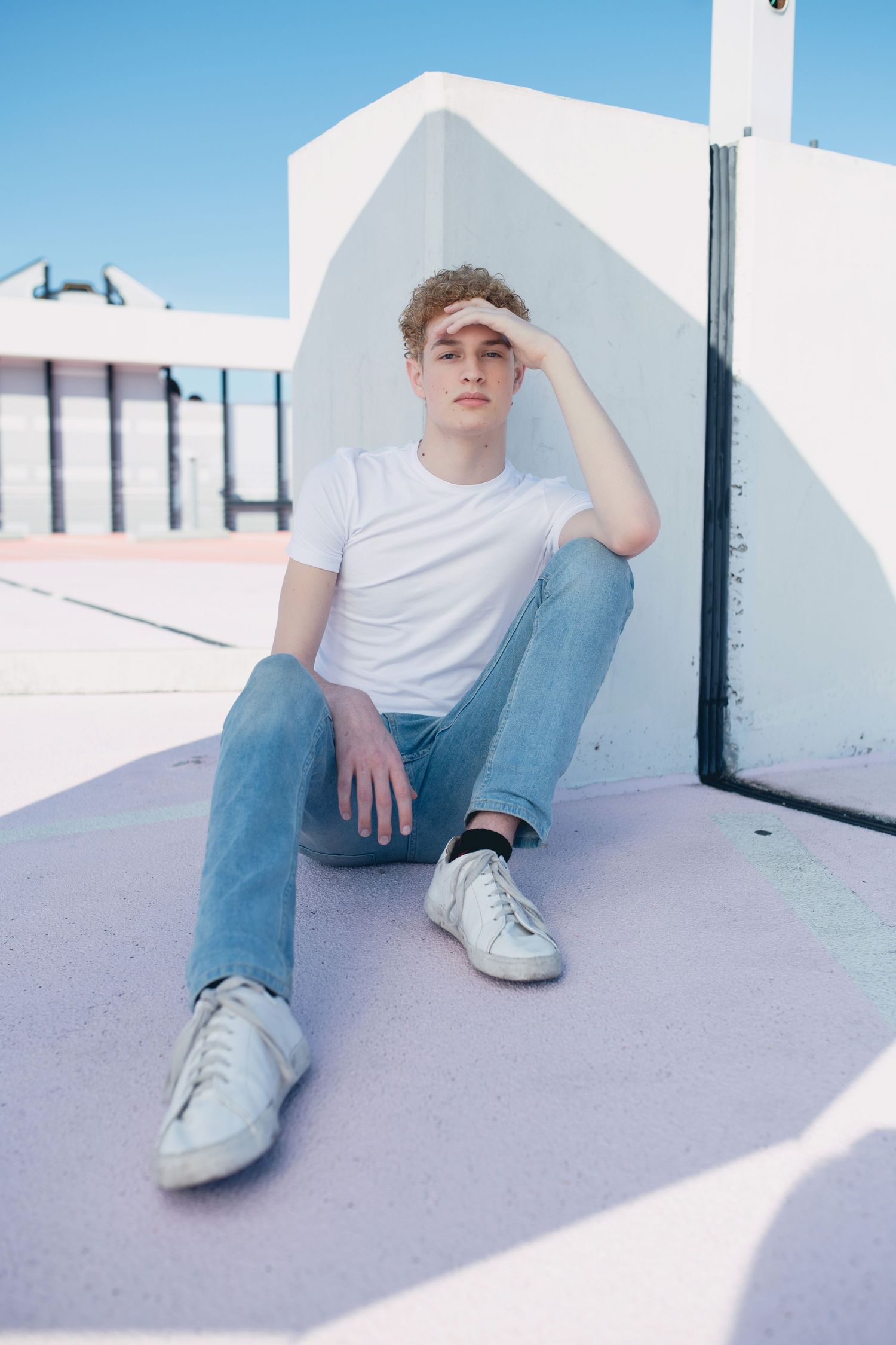 Young man in white t-shirt and jeans sitting on a rooftop under a bright blue sky.
