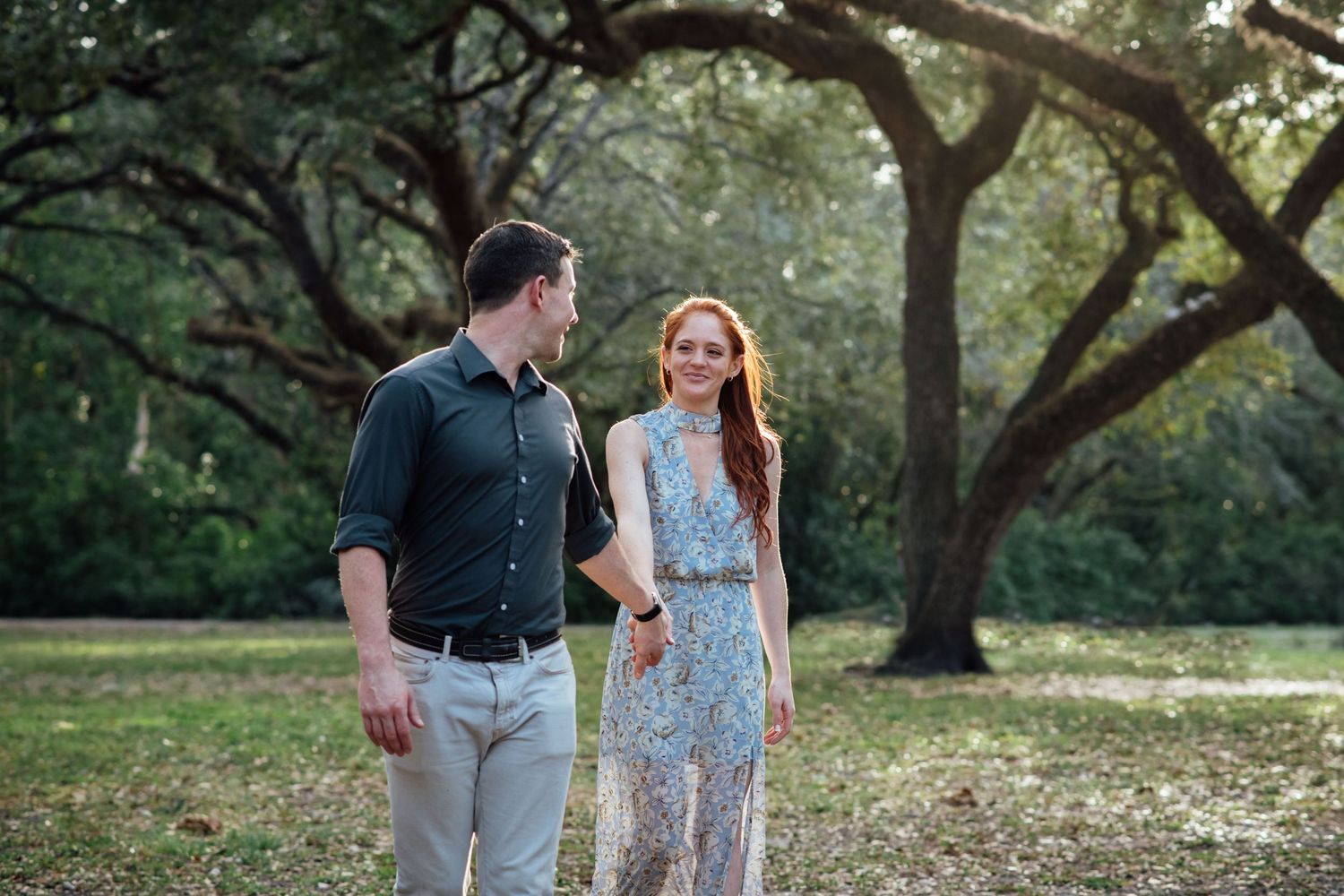 Couple walking together in a sunlit park with large oak trees, woman in floral dress smiling at man in dark shirt.