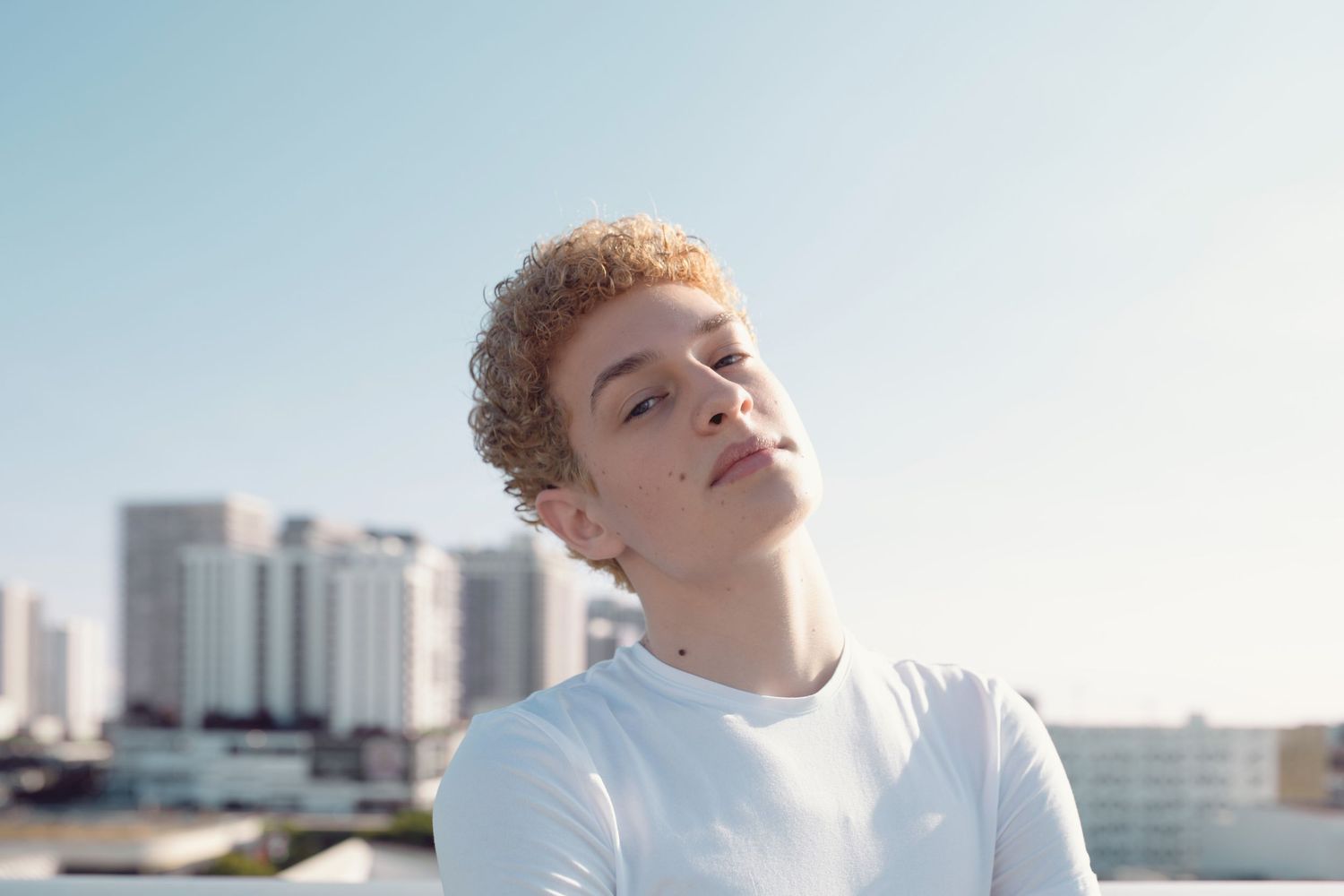 Young man with short curly red hair in a white shirt posing outdoors with a city skyline.