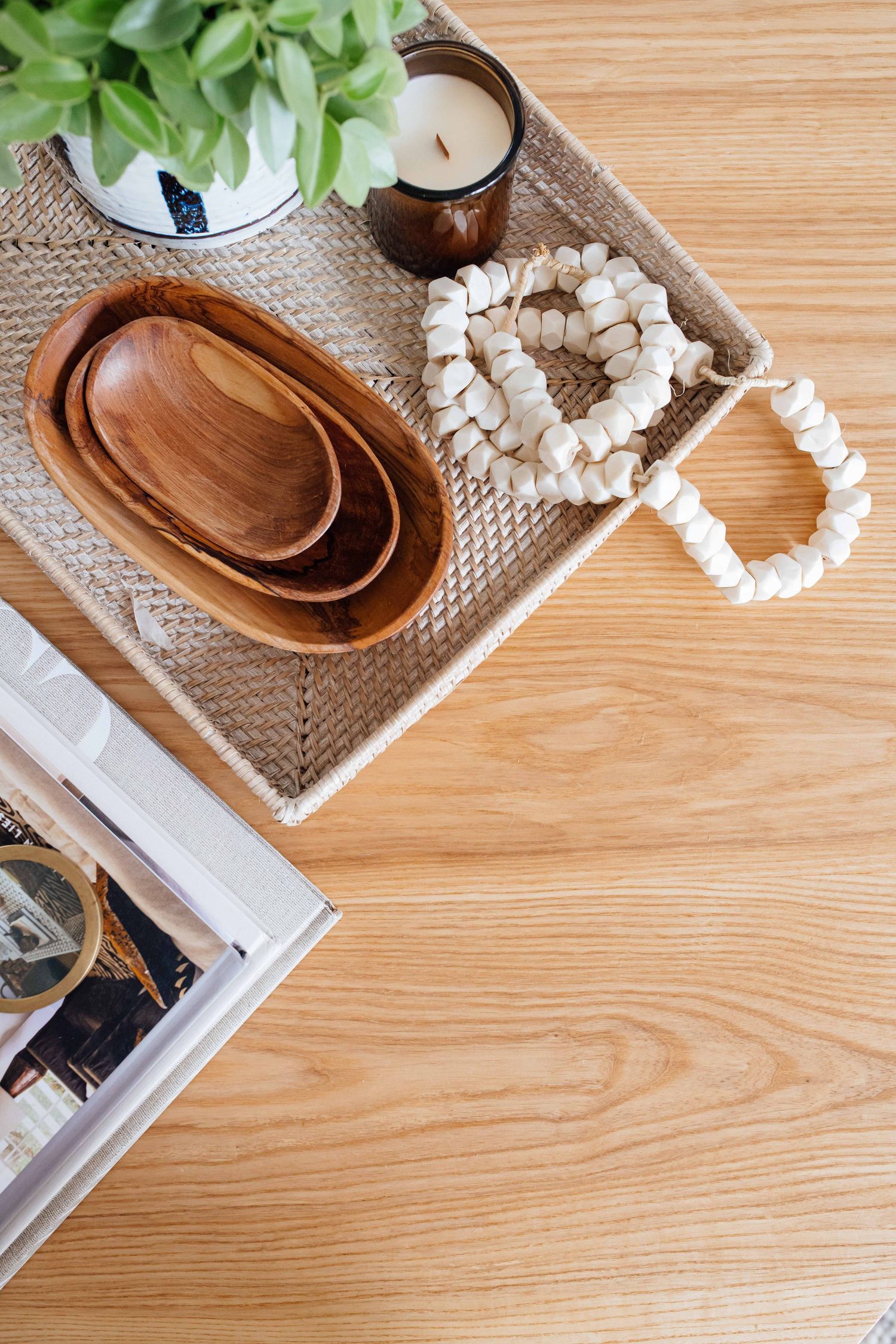 Wooden bowls, white bead garland, and candle styled on woven tray atop light wood coffee table.