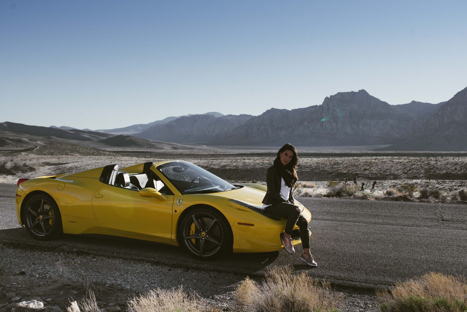 Woman leaning on yellow Ferrari convertible on desert highway with mountains.