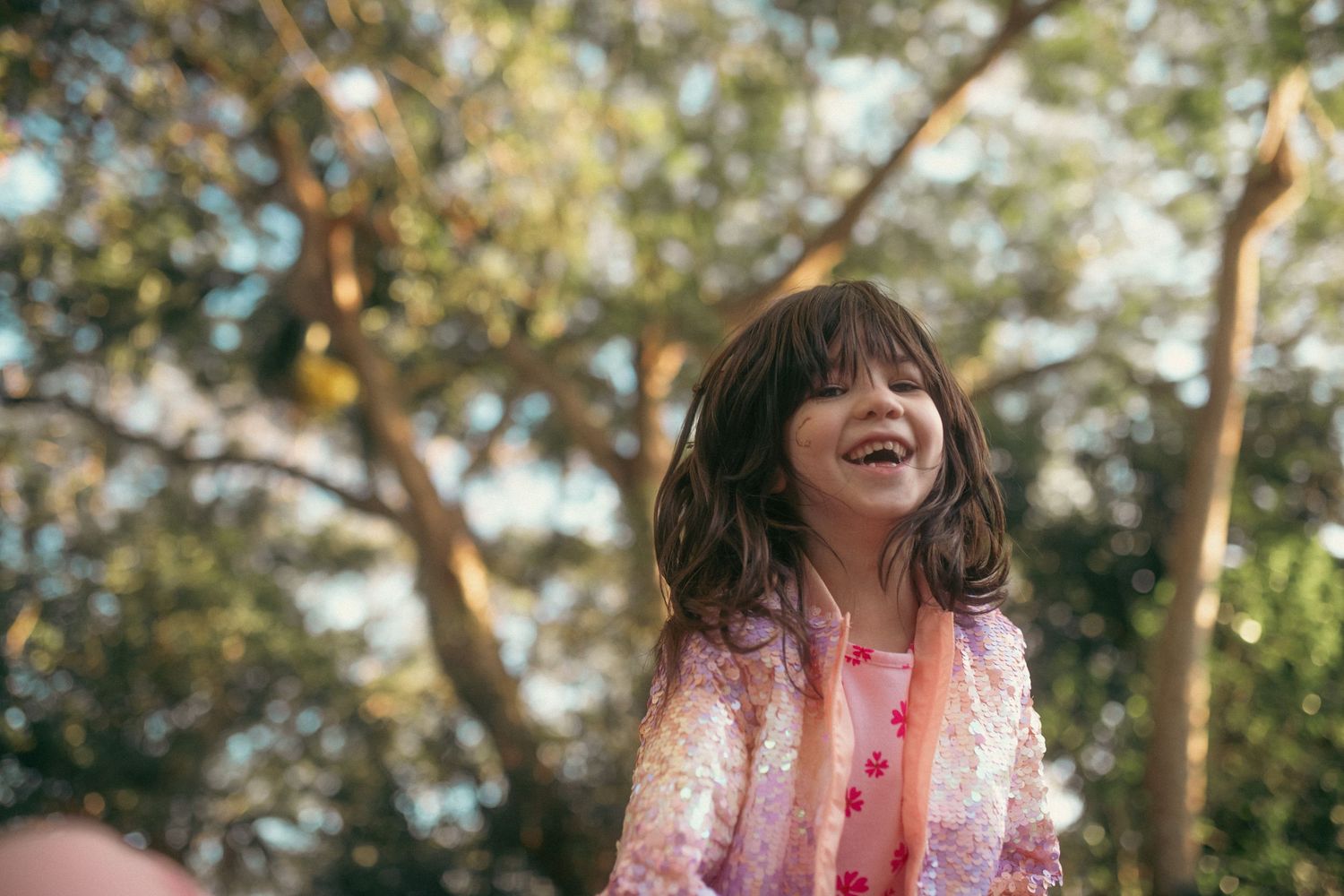 Joyful young girl laughing outdoors in pink sequin jacket surrounded by lush green trees.