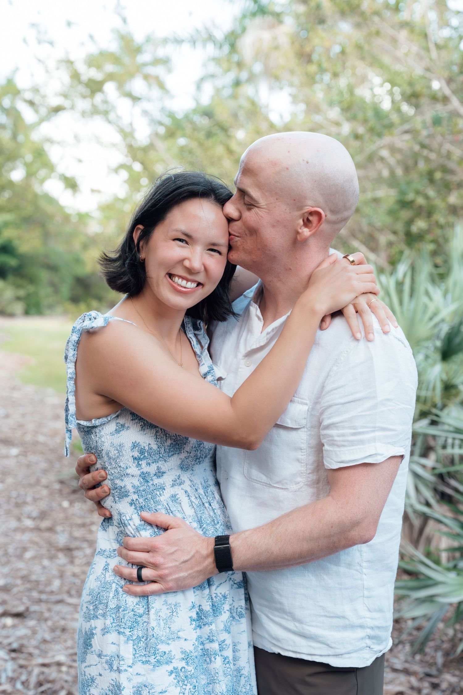A bald man kisses a smiling woman in a blue floral dress outdoors, both embracing warmly among green trees.