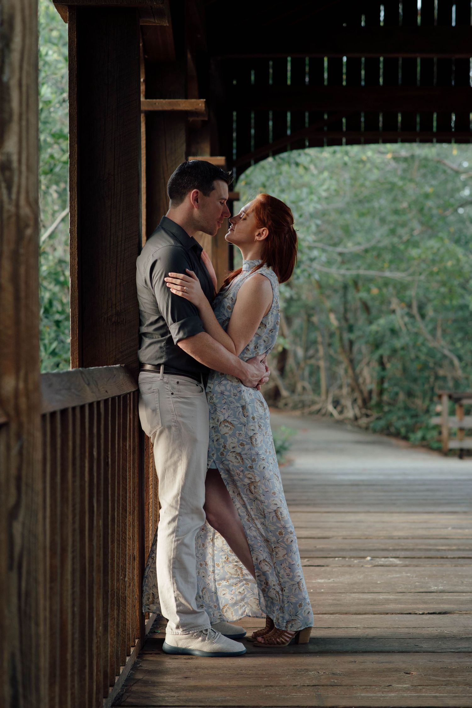 Couple sharing a romantic kiss on a wooden covered bridge walkway surrounded by lush green trees.
