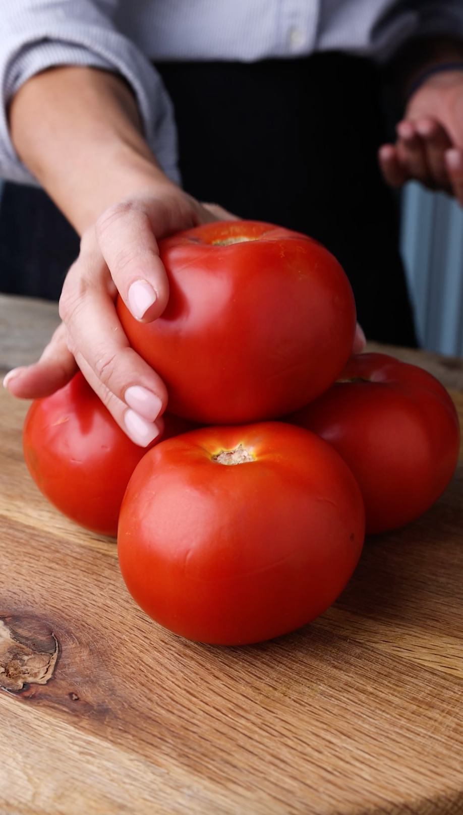 Person holding fresh red tomatoes stacked on a wooden cutting board for food photography.