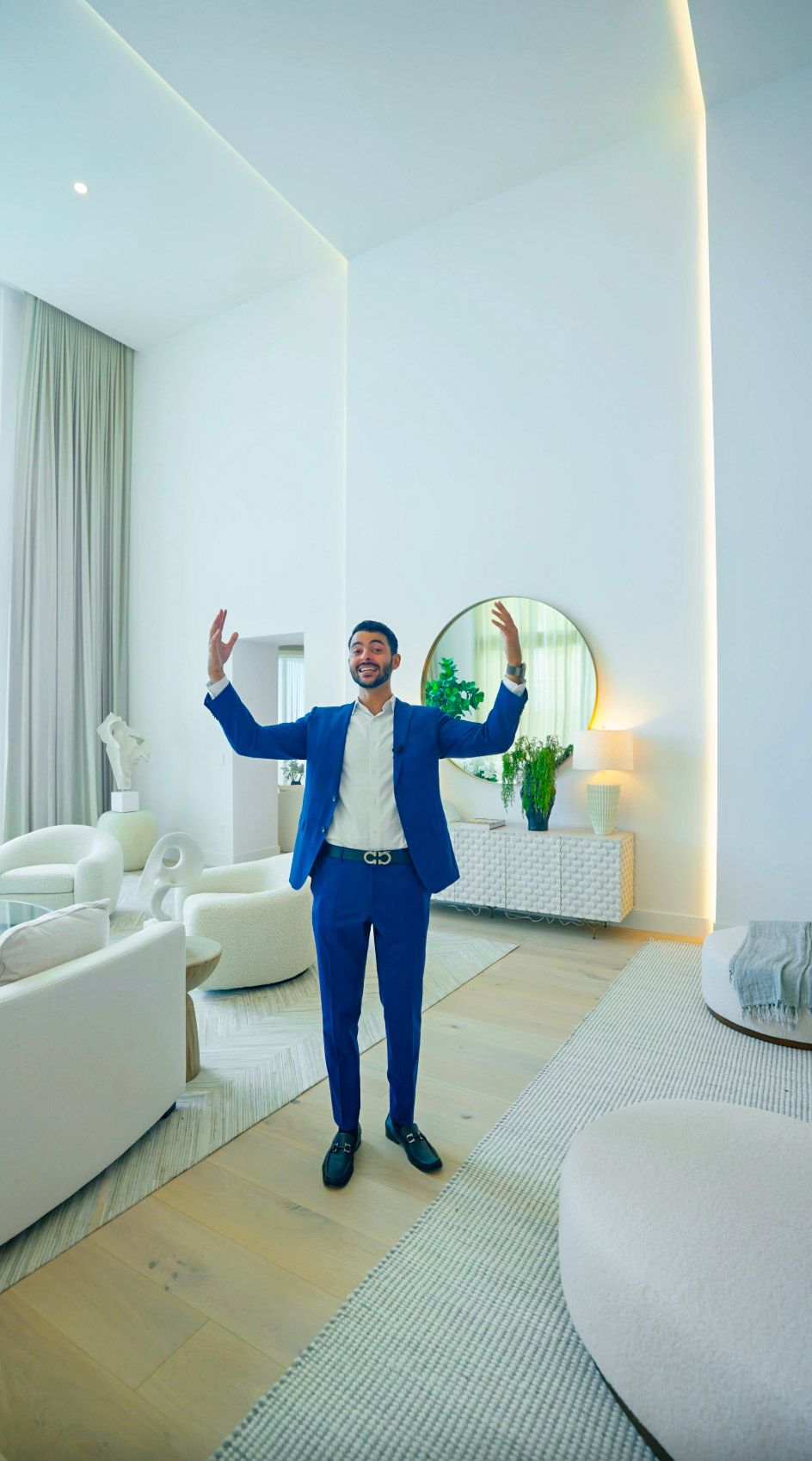 A person wearing a bright blue suit stands with raised arms in a modern white interior room with circular mirror decor.