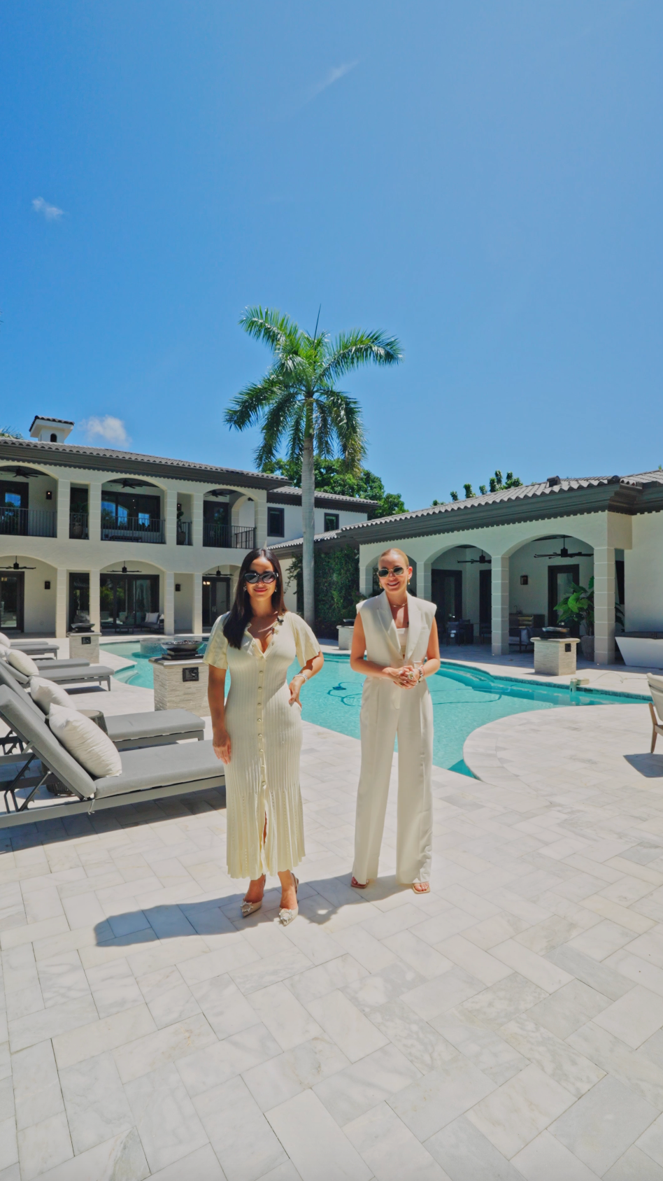 Two people in cream colored outfits stand on a patio beside a luxury swimming pool with palm trees and mansion in background.