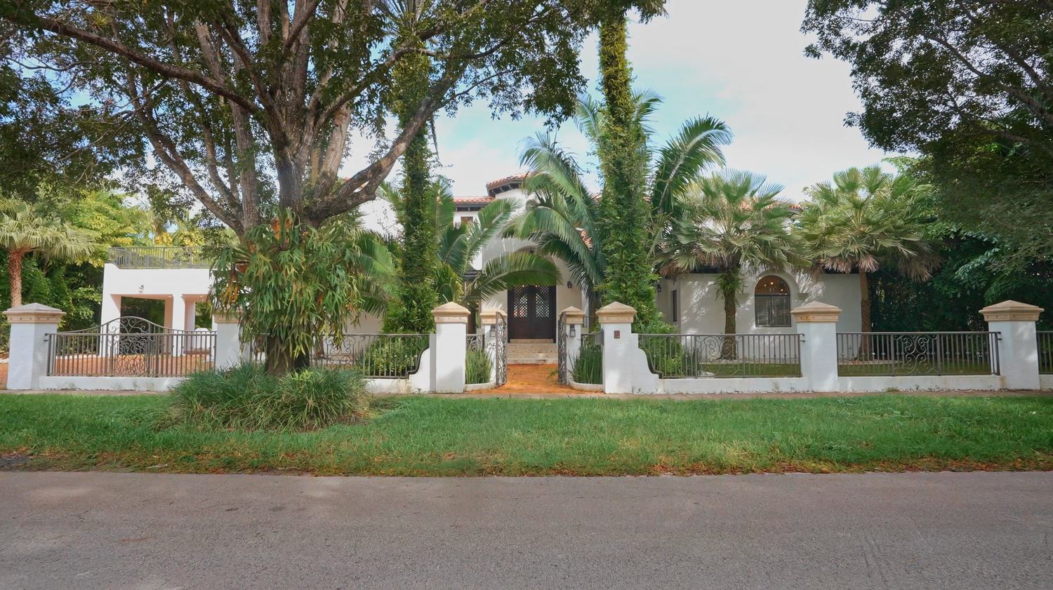 White fence surrounds a Mediterranean style home with lush palm trees and landscaping along a residential street.