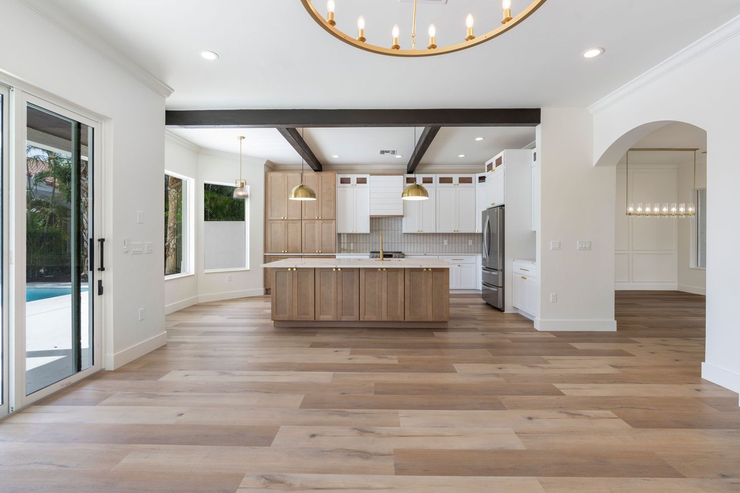 Modern open concept kitchen with light wood cabinets, exposed beams, and wide plank flooring opens to a poolside patio door.