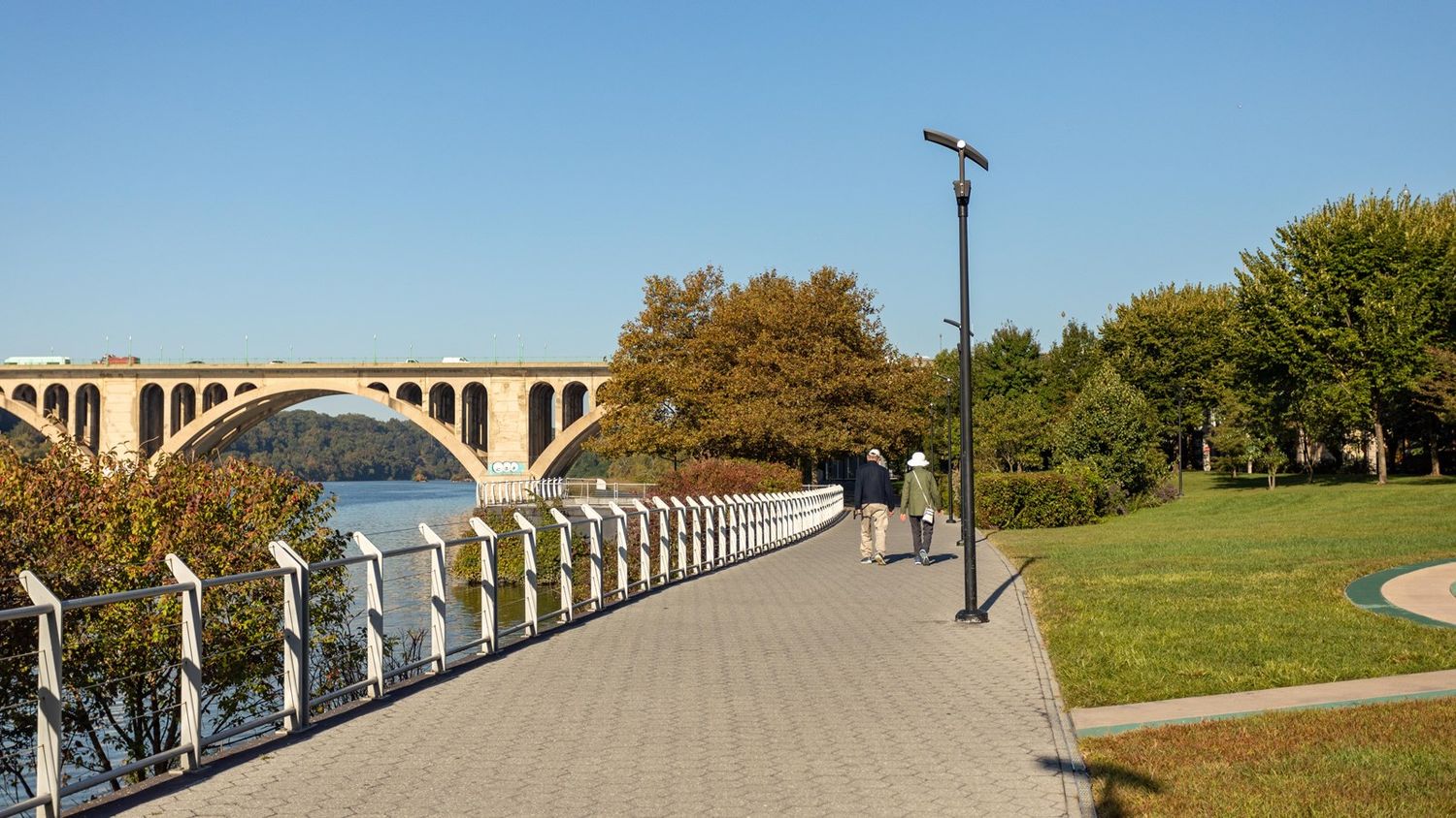 A tranquil riverside trail with walking path and bridge stretches along the water under a bright blue sky.
