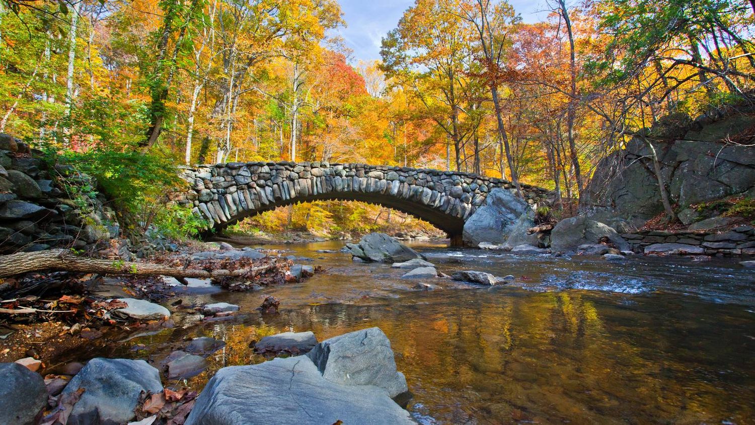 Stone bridge arch spans a tranquil stream surrounded by vibrant autumn foliage and rocky riverbed in a forest setting.