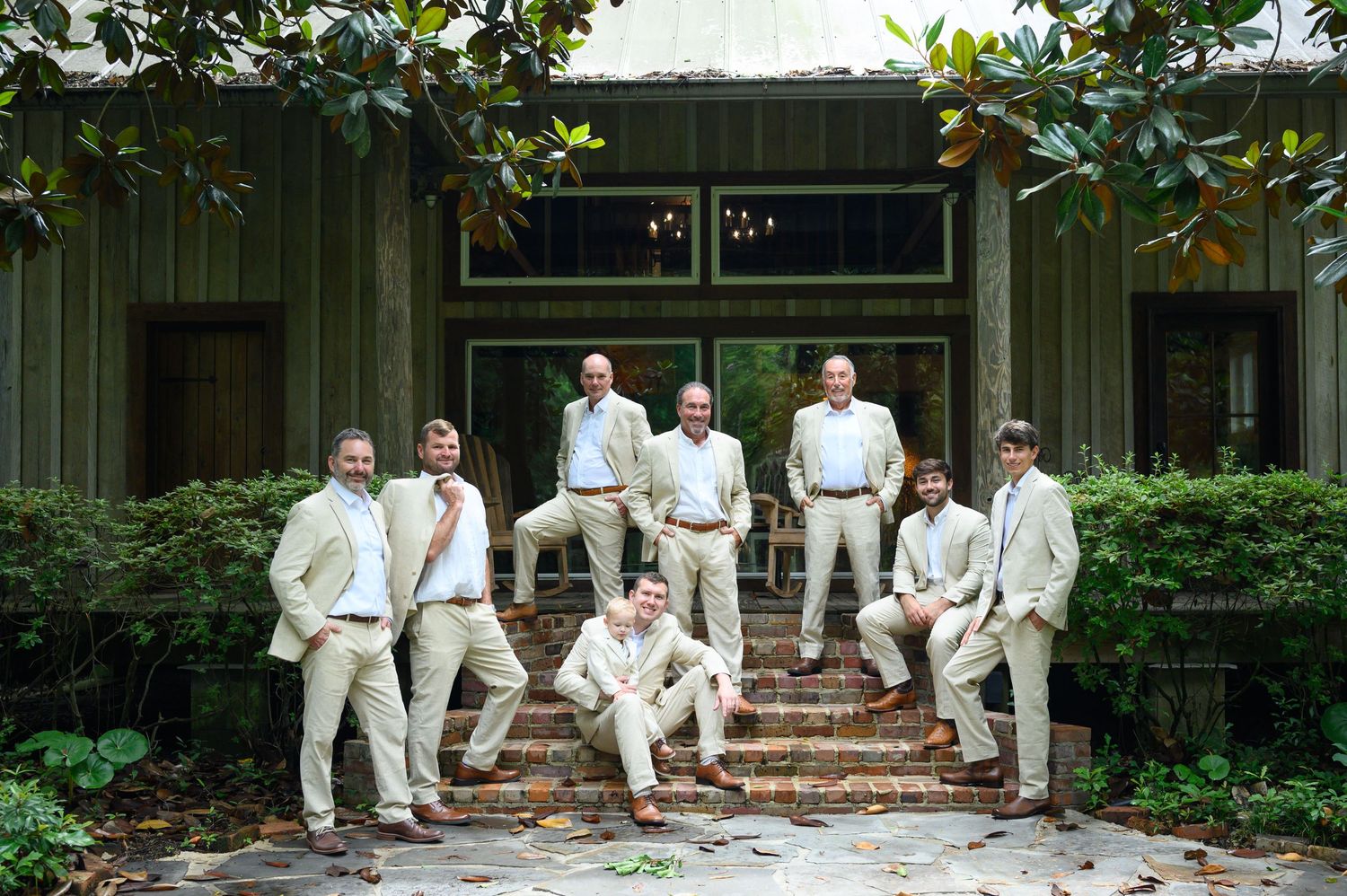 Groomsmen in matching tan suits pose on brick steps outside a rustic venue surrounded by lush greenery.