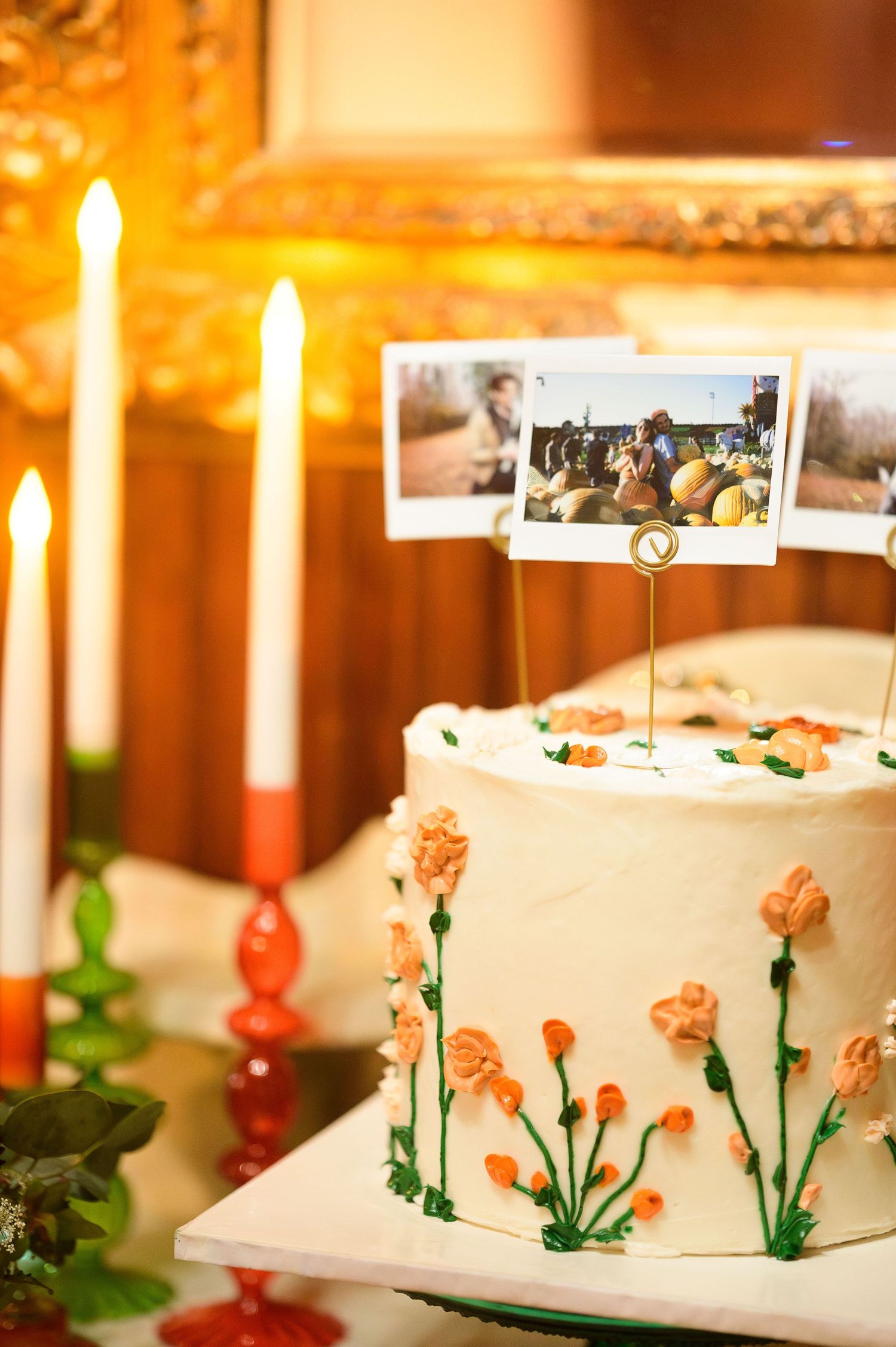 Wedding cake decorated with orange flowers and polaroid photos, surrounded by lit candles.