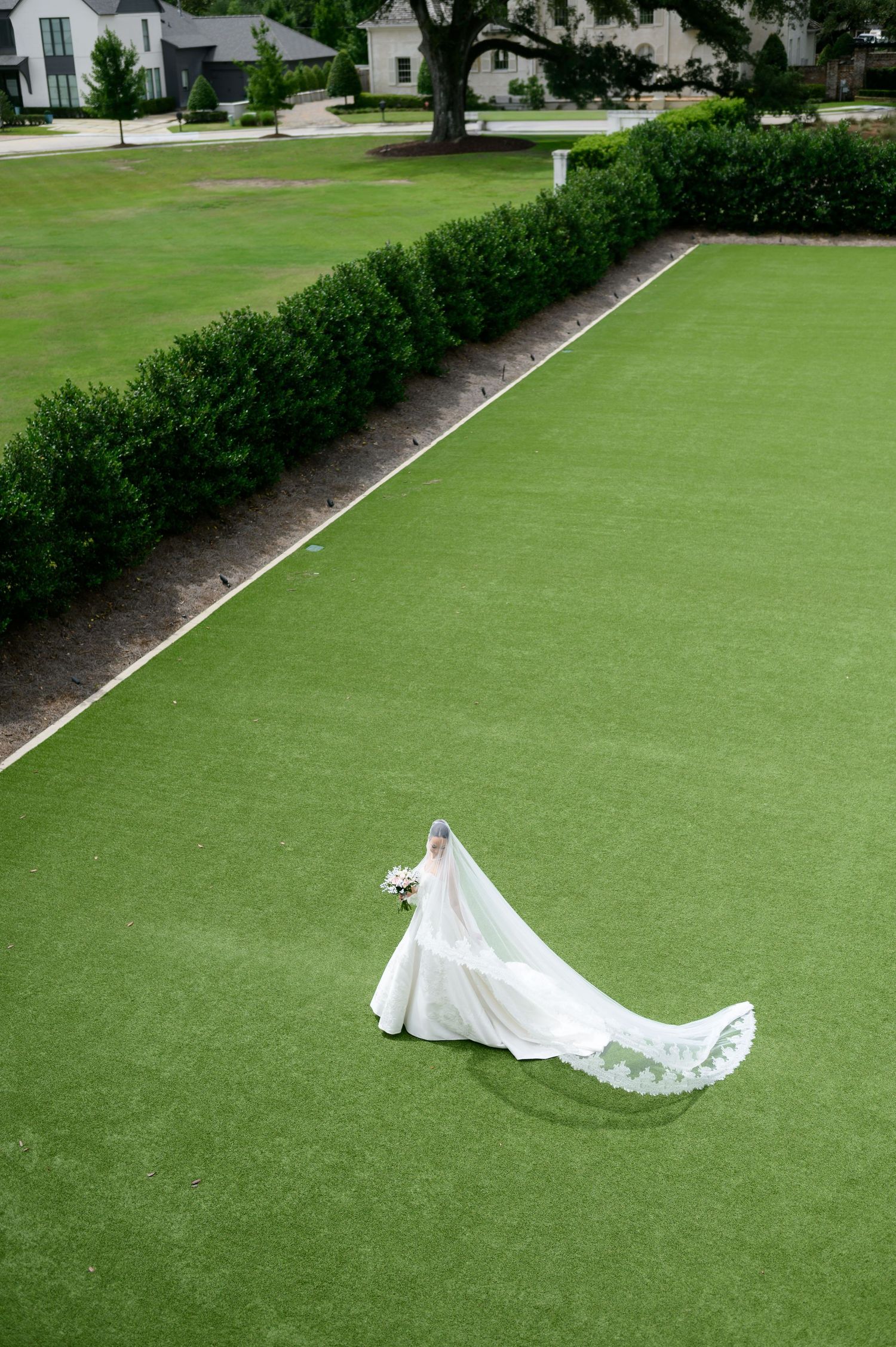 Aerial view of a bride in a white gown with long train and veil walking across a lush green lawn.
