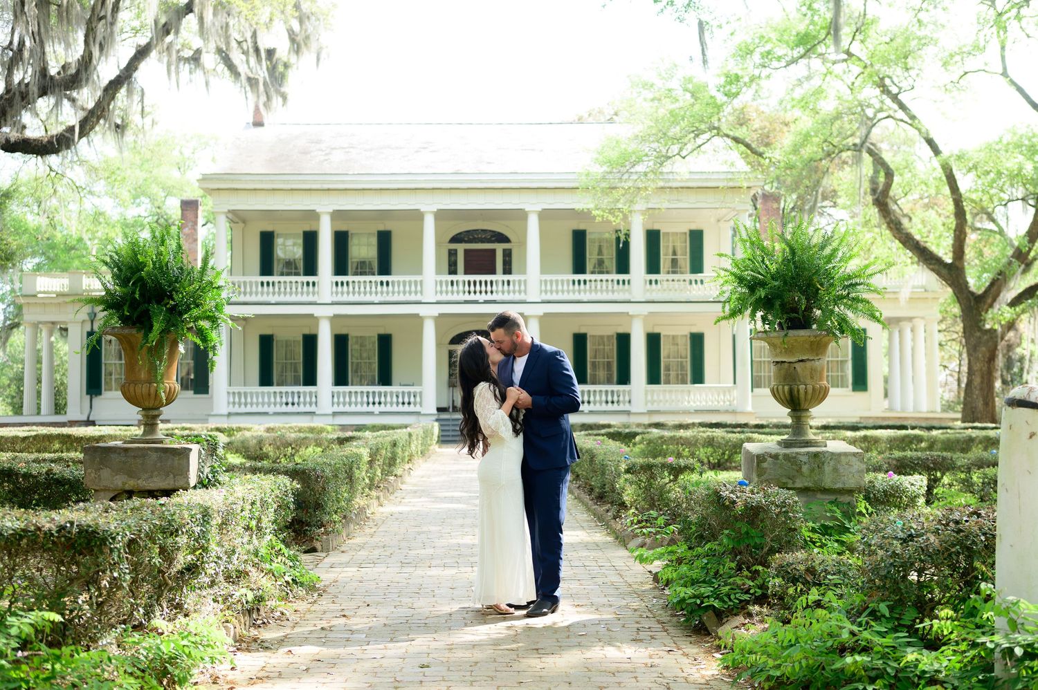 Bride and groom share a romantic kiss on a garden path in front of a grand Southern plantation mansion.