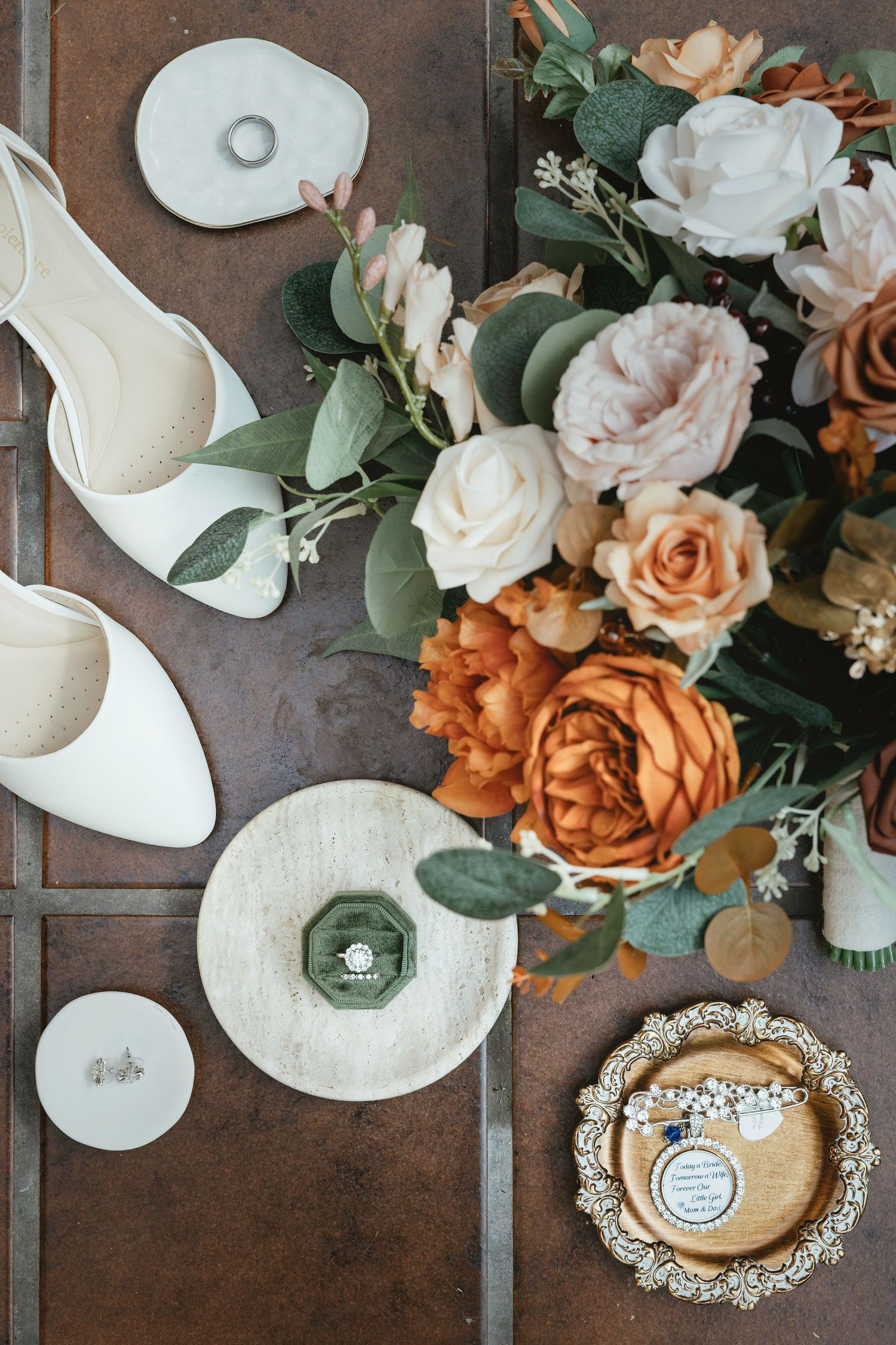Wedding accessories including white shoes, rings, and a bouquet of peach and cream roses with eucalyptus arranged on dark surface.
