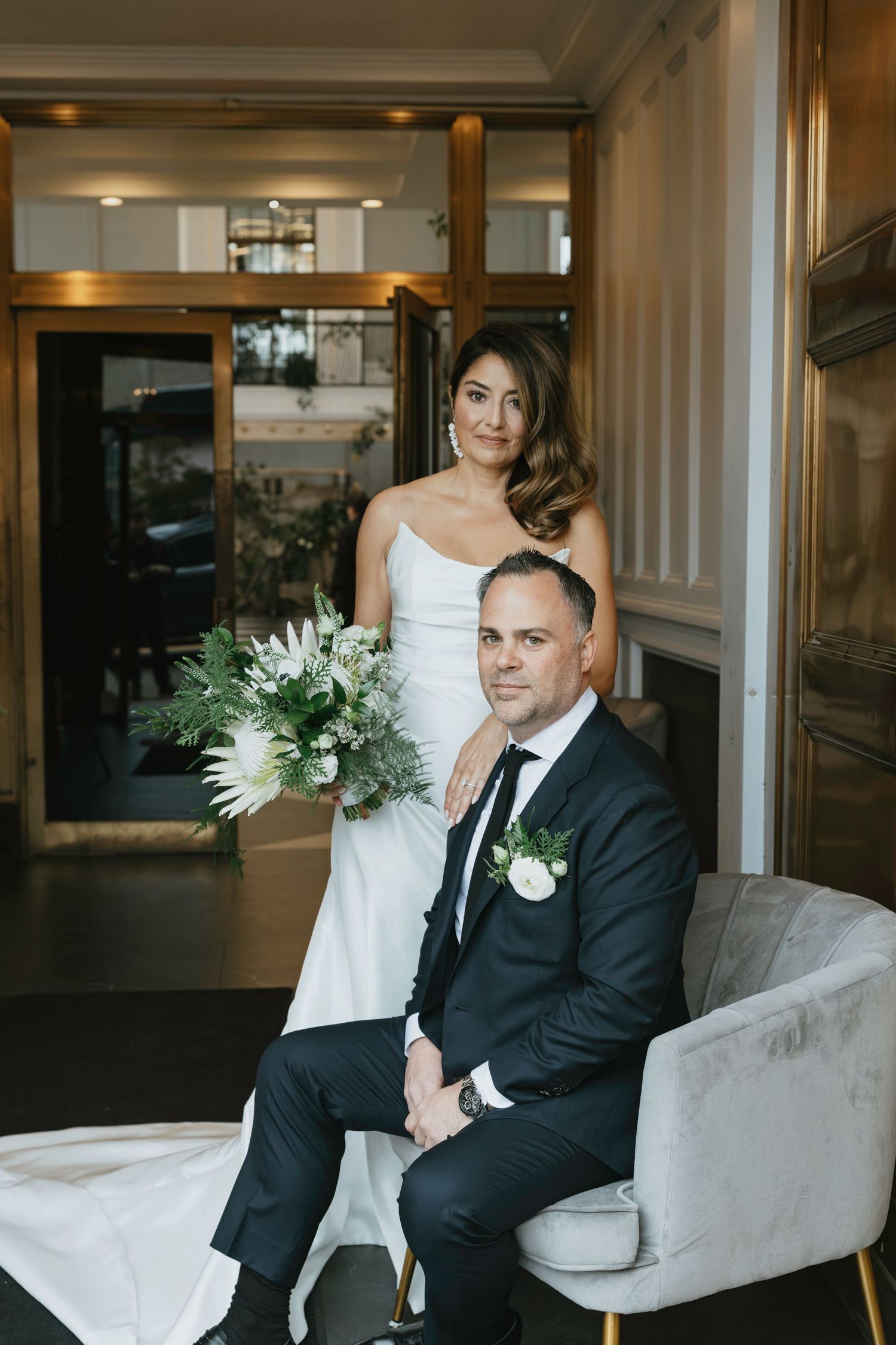 A wedding couple sits together on a light gray couch in an elegant indoor setting with white floral arrangements.