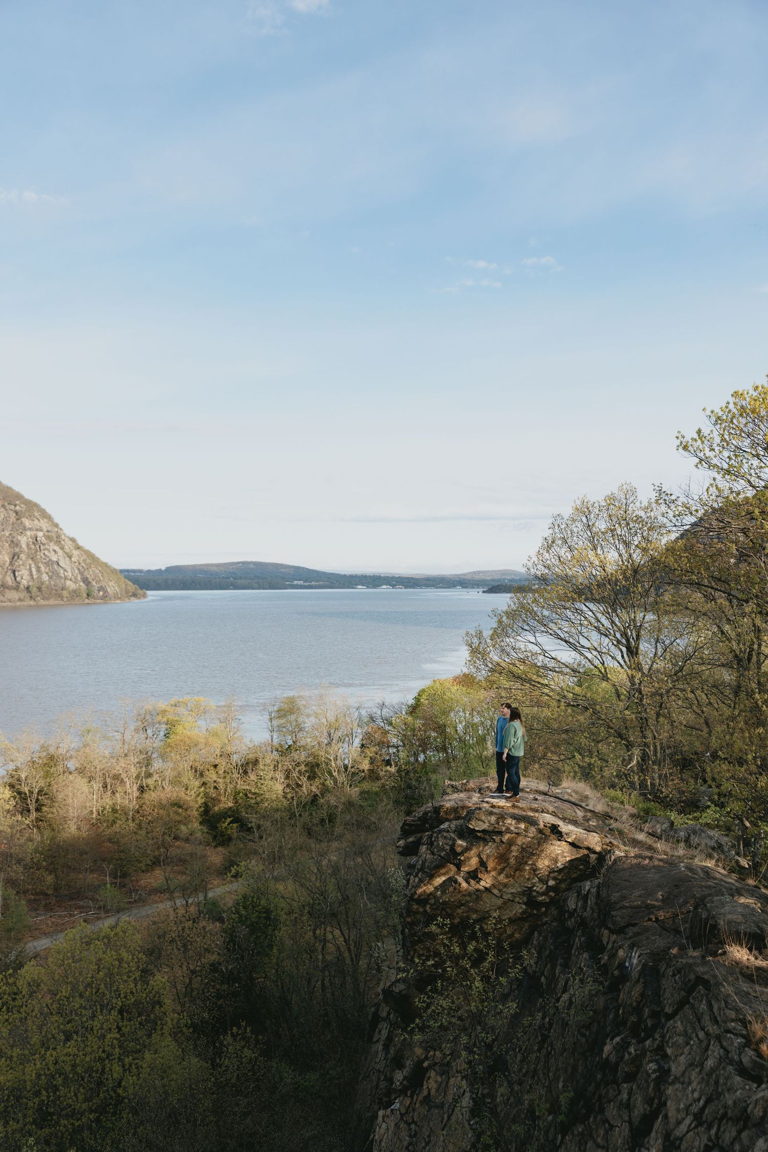 A hiker stands on a rocky overlook above lush trees with a wide river and distant mountains under a blue sky.