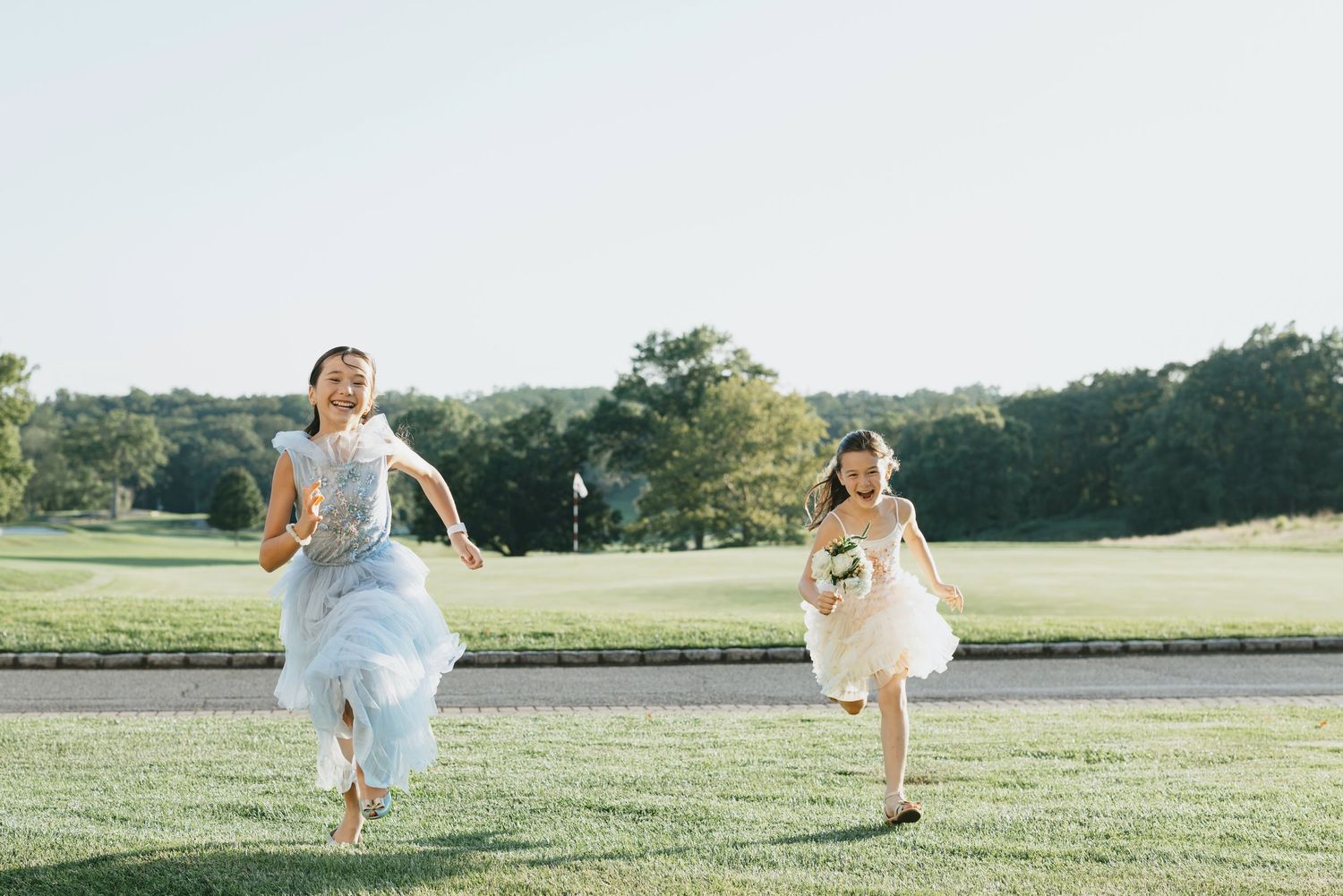 Wedding party running and twirling in white dresses across a sunny green lawn.