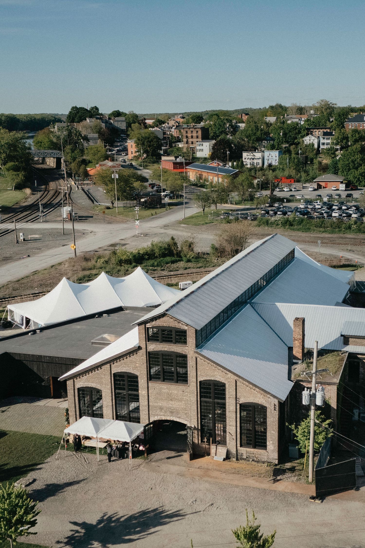 Aerial view of a historic brick industrial building with white tent structures near railroad tracks in a small town.