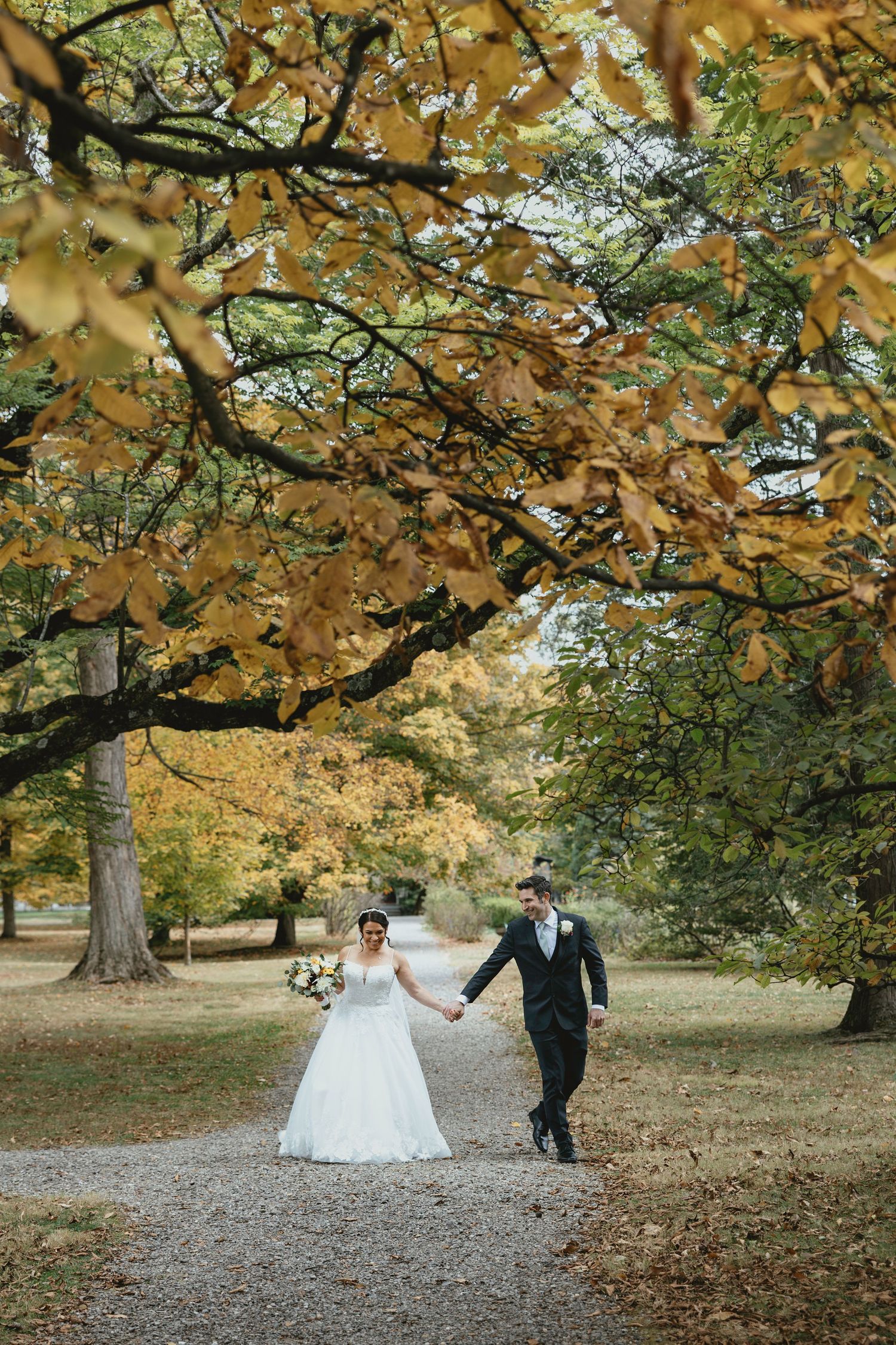 A couple walks through a park with golden autumn leaves overhead during their wedding photo session.
