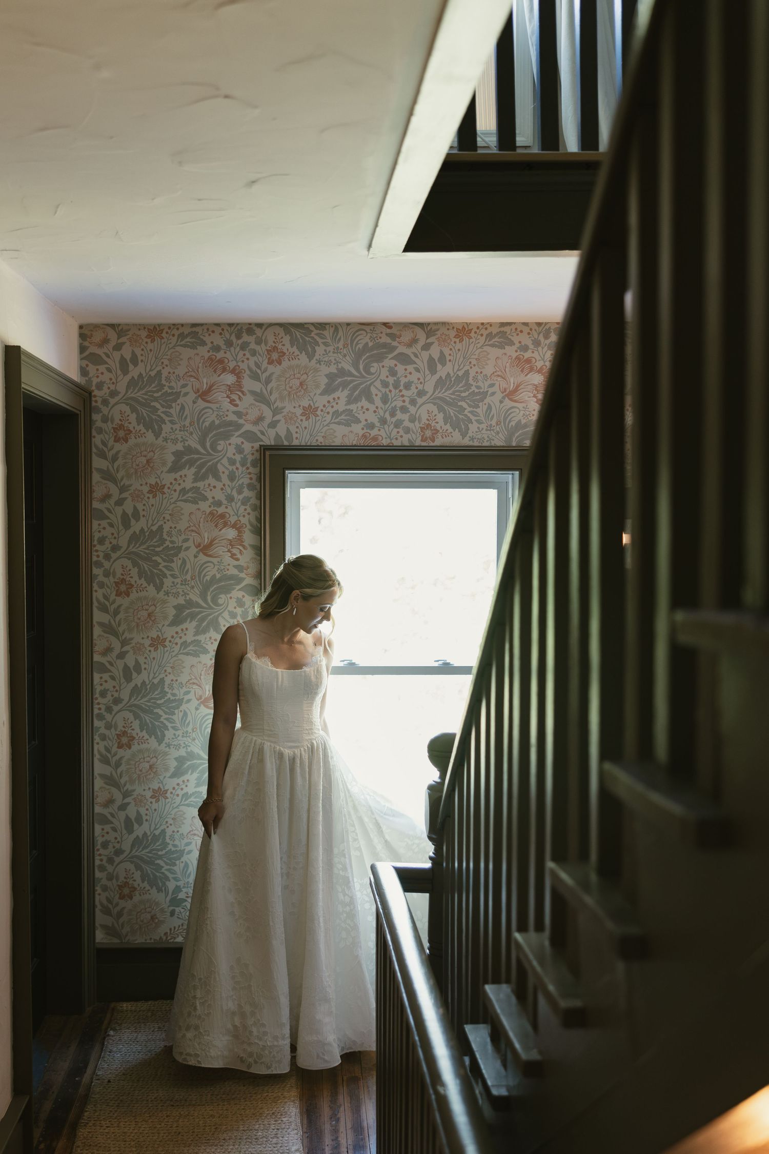 Bride in white wedding gown standing by window on staircase landing with floral wallpaper in vintage home.