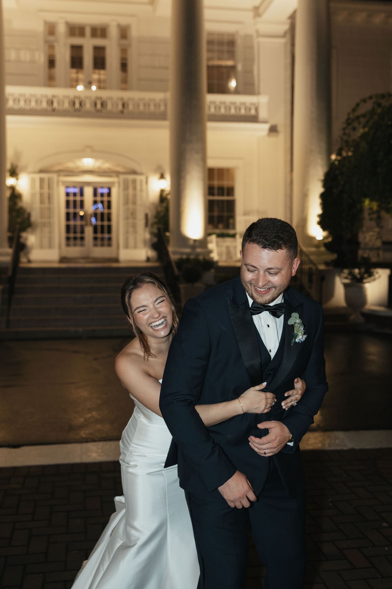 Bride in white gown laughing joyfully while embracing groom in navy tuxedo outside illuminated wedding venue at night.