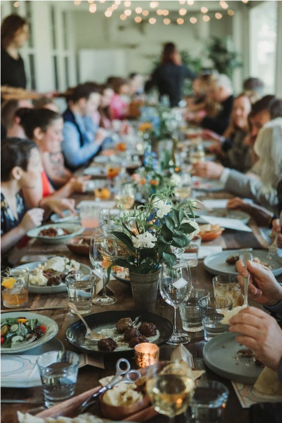 Guests dining at a long wooden table with floral centerpieces, wine glasses, and plates of food at an elegant event.