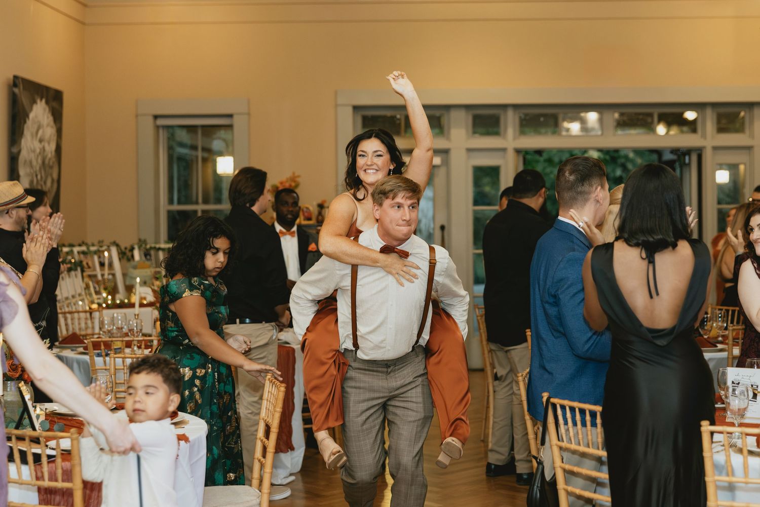 Guests dancing and celebrating at an indoor wedding reception with warm lighting and wooden chairs.