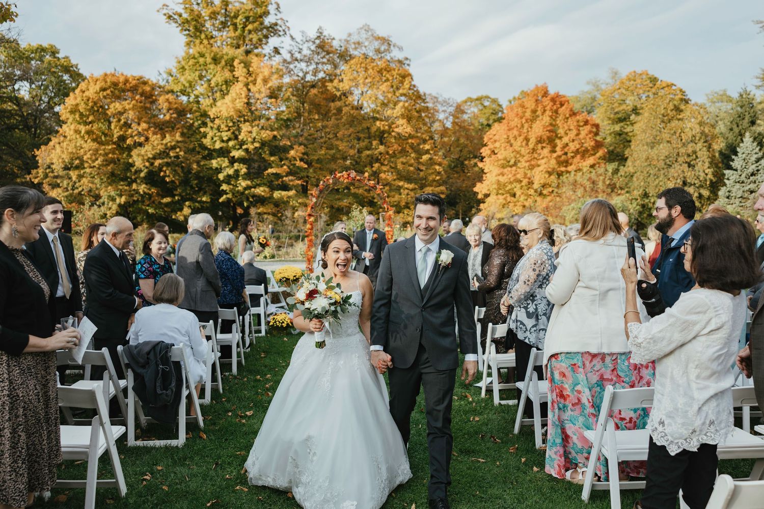 A wedding ceremony takes place outdoors on a beautiful autumn day with golden foliage and white chairs set up on the lawn.