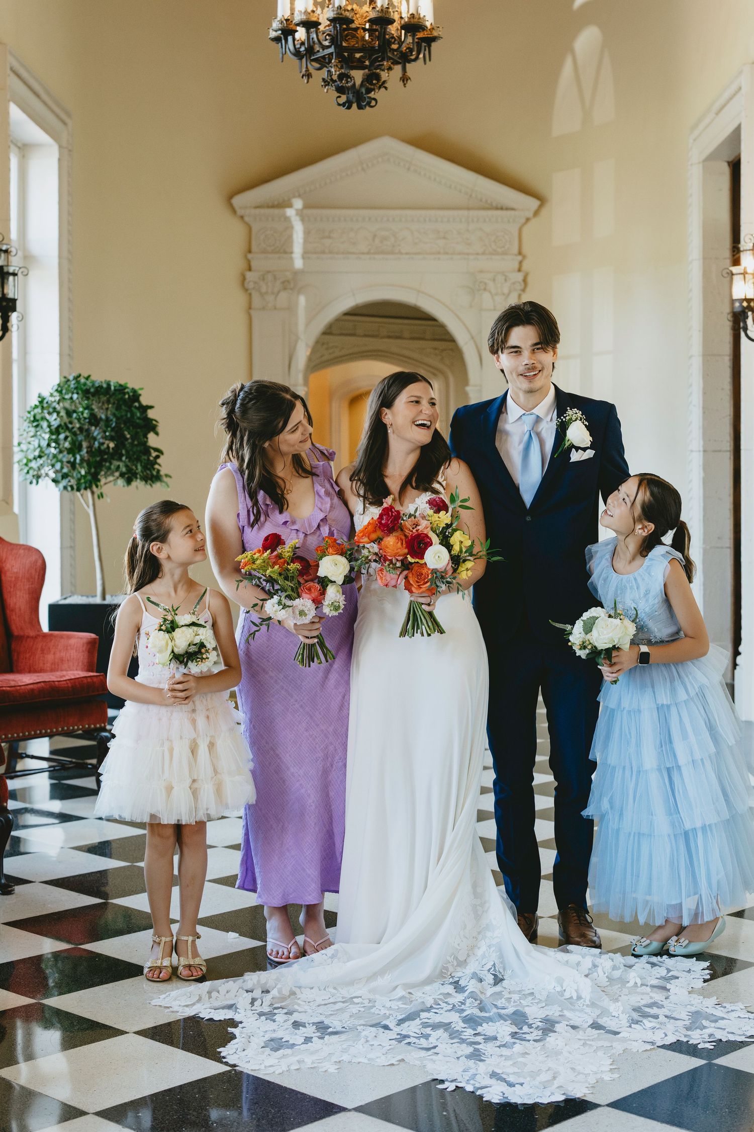 A wedding party gathers on a black and white checkered floor in an elegant room with chandeliers.